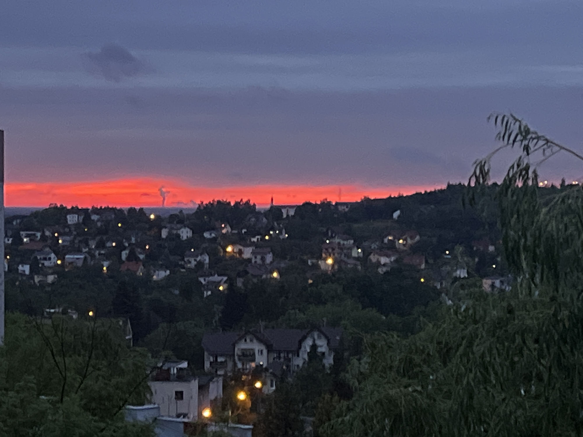 An exterior, eye-level, wide shot captures a vibrant, ruddy sunset over a densely populated, hilly urban area. The sky transitions from a deep, dusky purple at the top of the frame to a fiery orange and red band just above the horizon, with wispy, dark clouds scattered across the purple expanse. Below the horizon, the landscape is dotted with numerous houses, some with illuminated windows, nestled among dark green trees. The houses appear to be of varying sizes and architectural styles, contributing to a textured, uneven cityscape. In the foreground, the dark, leafy branches of trees obscure parts of the lower section of the image, adding depth and a sense of being within a natural environment overlooking the city. The overall scene suggests a residential area during twilight, with the dramatic sunset providing a striking backdrop.