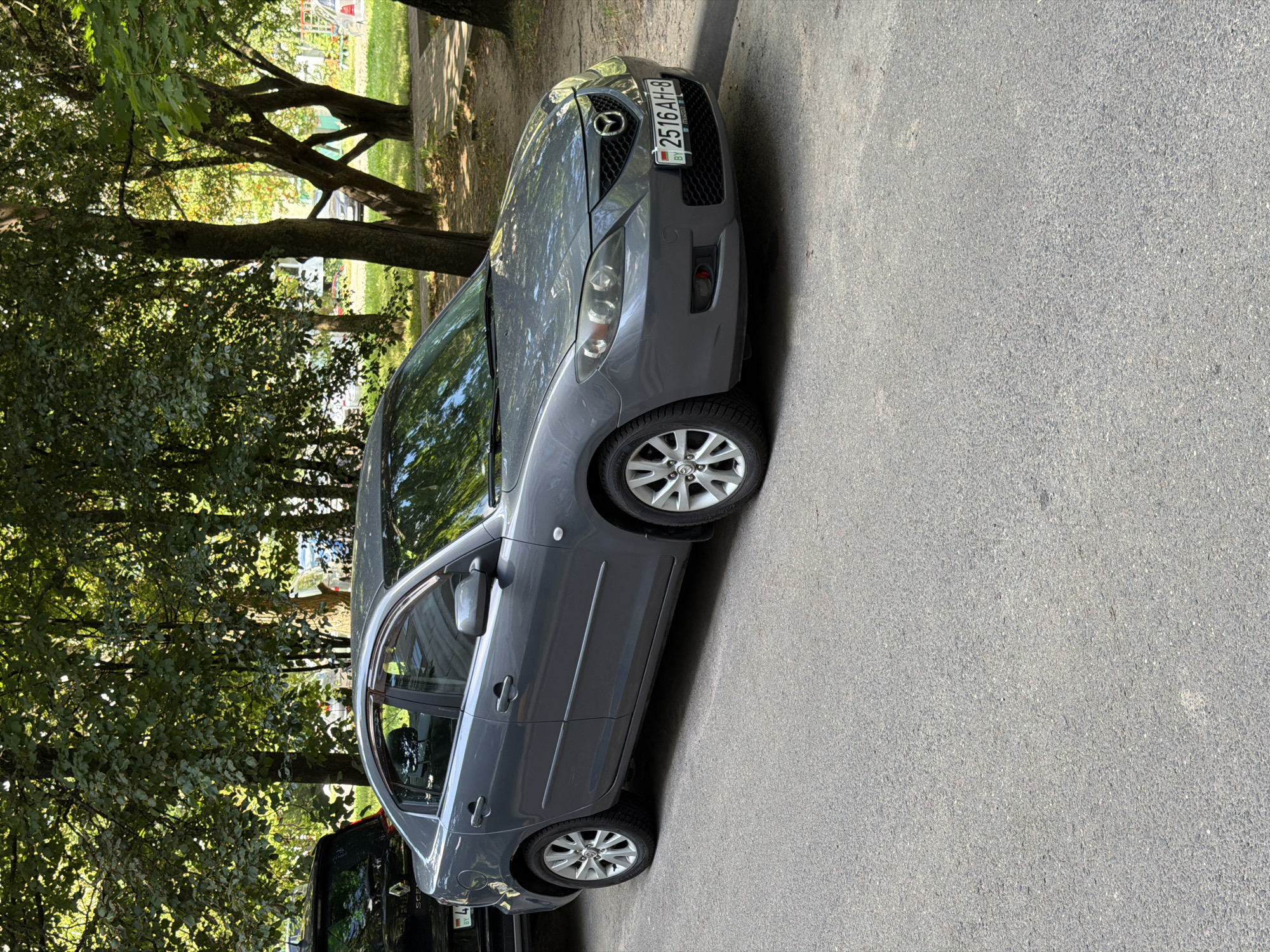 A grey Mazda sedan is parked on a paved road in a city, with trees and grass visible in the background. The car has a license plate that reads "2516 AH-8". The scene appears to be in Minsk, Belarus, as indicated by the "BY" on the license plate, which is the country code for Belarus. The car is positioned in the foreground, with its front end facing slightly to the right. The sun is shining, casting shadows from the trees onto the car and the road. There is another car parked behind the Mazda, a black SUV. The overall environment suggests a typical urban setting, possibly a residential area or a park. The image is taken from a slightly elevated angle, looking down at the car. There are no people visible in the image.