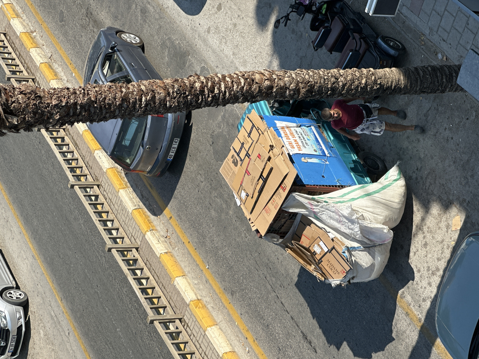 This is an overhead, high-angle shot of a street scene, possibly in Fethiye, Turkey. A prominent palm tree trunk runs diagonally across the frame, partially obscuring the view. On the right side of the image, a man in a red shirt and patterned shorts is lying on the ground next to a blue vehicle loaded with cardboard boxes. One of the boxes has text on it, including "MADAME COCO" and "GERİ DÖNSÜM YAŞAM SÜRSÜ LÜTFEN İÇİNE ATINIZI ÇÖP ATMAYINIZ! FETHIYE," which translates to "Recycling Life is a Surprise Please put it inside Do not throw trash! Fethiye." This suggests the vehicle is related to recycling or waste collection.
To the left of the palm tree, a dark gray car is parked on the side of the road. The road has yellow lane markings and a raised curb with what appears to be a wooden structure resembling a staircase or ramp. Further to the left, the front of a silver car is visible. A dark-colored scooter is parked behind the blue vehicle on the right. The scene appears to be set in daylight, with strong shadows cast by the palm tree and other objects, indicating a sunny day. The overall atmosphere is utilitarian, possibly depicting a moment of rest or work activity on a busy street.
