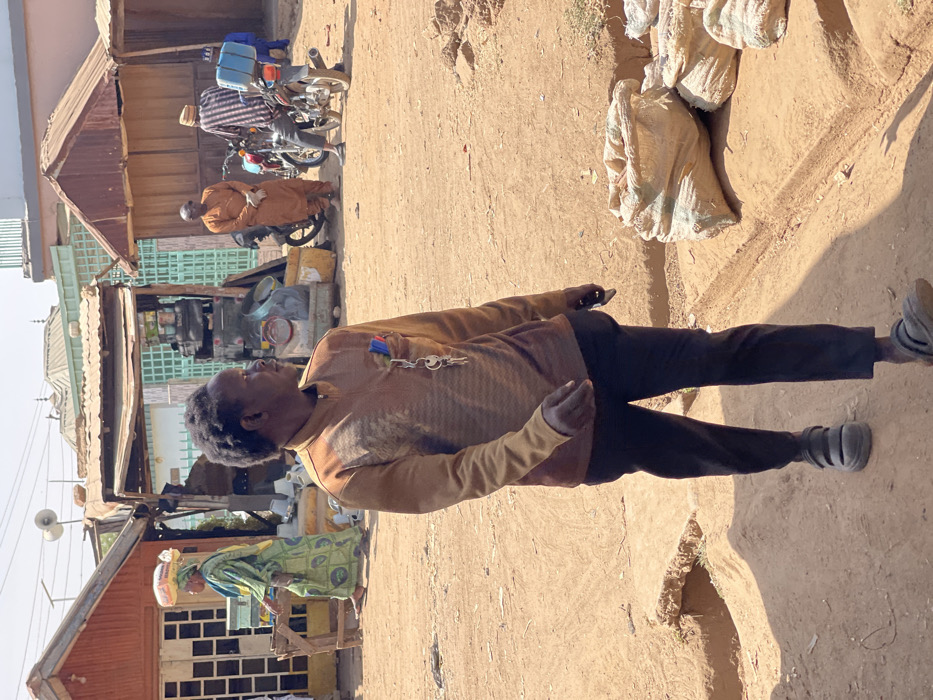 The scene depicts an unpaved commercial area in Chikaji, Nigeria, under daylight conditions. In the foreground, a dark-skinned male, wearing a brown patterned long-sleeved shirt, dark pants, and dark sandals, walks rightward. A pen and keys are clipped to his left chest pocket. To his right, several beige sacks are piled on an elevated dirt section. In the midground, a woman in a green and yellow dress with a white headwrap carries a white sack on her head, moving left past a reddish-brown wooden stall. Further right, two men are present near motorcycles: one in an orange garment stands next to a red motorcycle, while another in a striped shirt and hat is near a motorcycle fitted with a blue delivery box. The background features various simple structures with corrugated metal roofs, including one with loudspeakers. The ground consists of dry, reddish-brown dirt.