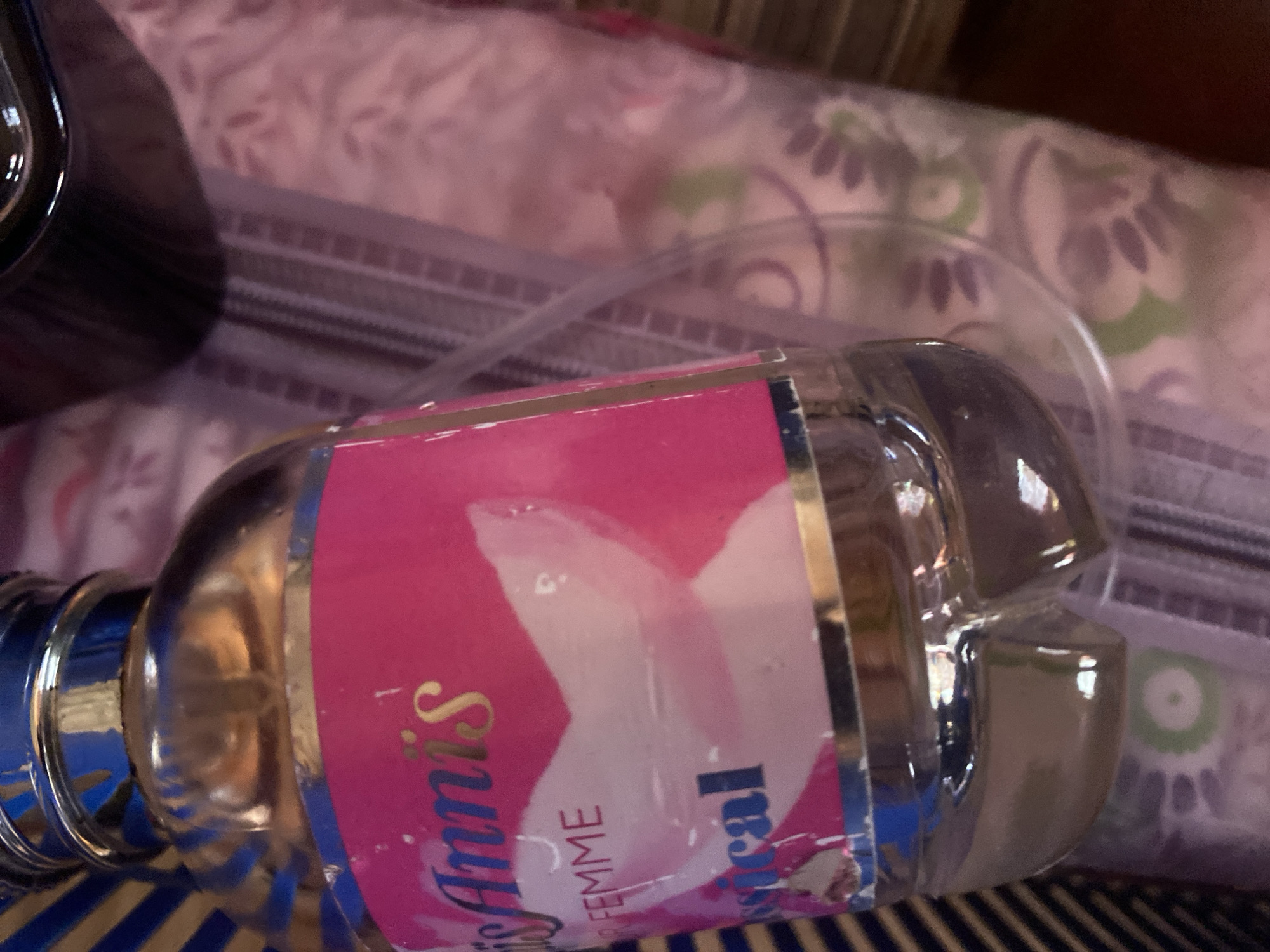 The image shows a close-up of a bottle of "Ann's Miracle Femme" perfume, lying on its side. The bottle is made of clear glass and has a shiny silver cap. The label is predominantly pink with white floral designs and gold lettering. Some of the text is visible: "Ann's," "MIRACLE," and "FEMME."
The bottle rests on a striped surface, likely a tablecloth or bedsheet, with alternating blue and dark blue stripes. In the background, a pink fabric with a subtle floral pattern is visible, suggesting an indoor setting, possibly a bedroom or living area. The lighting is soft and diffused, indicating it's likely daytime, but it's difficult to determine the exact time. There are no people visible in the image, and the focus is solely on the perfume bottle. The overall impression is one of domestic comfort, with the perfume bottle presented as a personal item.