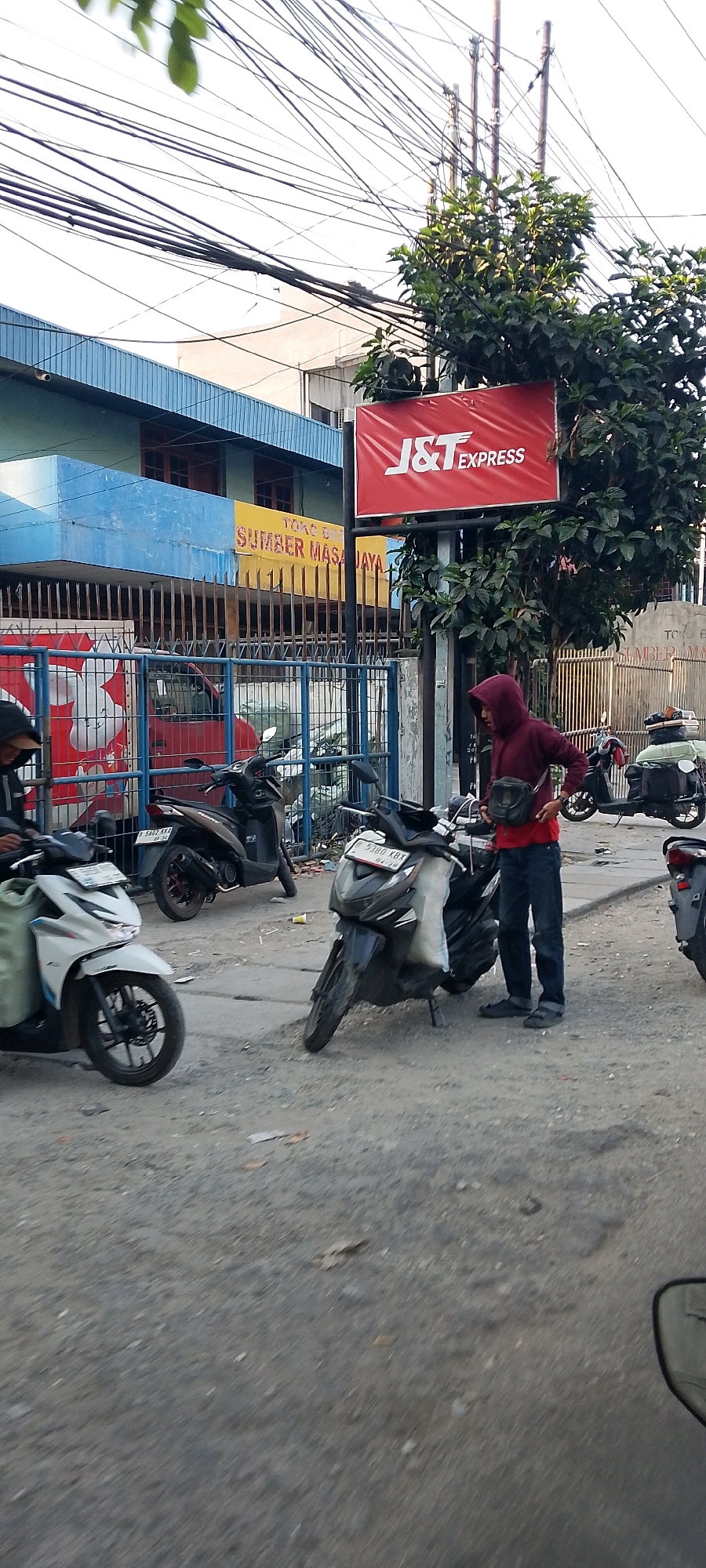 In Bekasi, Indonesia, a man wearing a red hoodie and dark pants stands next to a dark-colored scooter on a paved street. The scooter has a white bag on the back and a license plate with the characters "B 5380 KBX". The man is holding his right hand near his waist. Behind him, another person is partially visible on a white scooter. In the background, a blue and yellow building is visible, with a red sign that reads "J&T EXPRESS" mounted on a pole. A metal fence separates the street from the building. The street is lined with several other parked scooters, and the ground is covered with gravel and some litter. Overhead, a complex network of electrical wires crisscrosses the sky. The scene appears to be an outdoor area, possibly a street or parking lot, in a commercial or residential part of the city.
