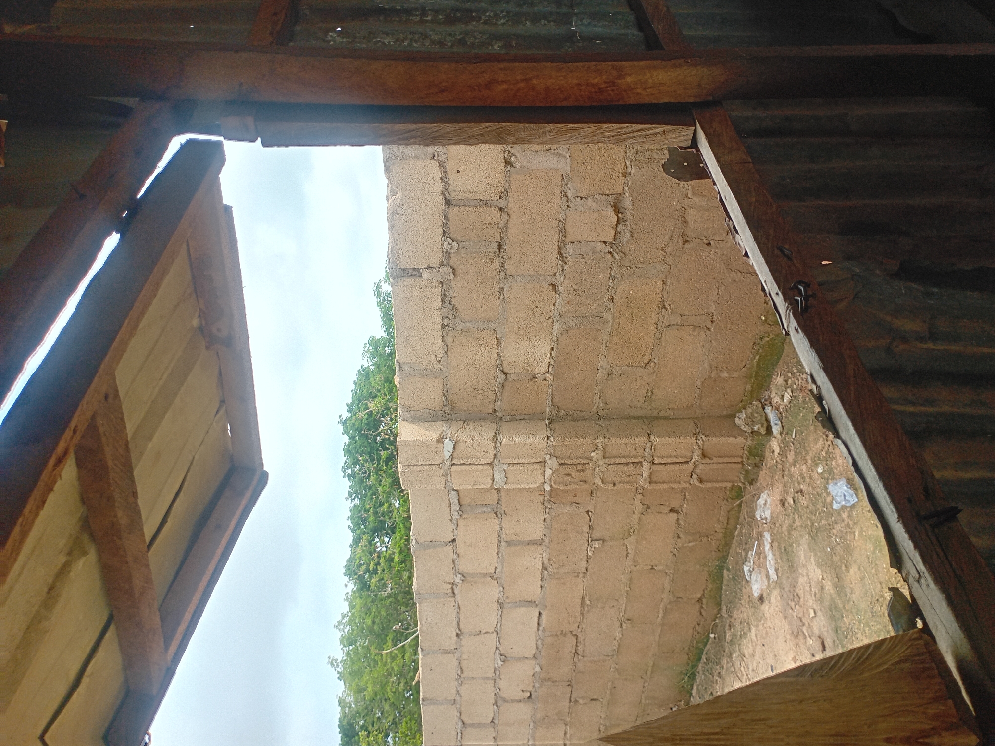 The image shows an outdoor scene from behind a partially constructed or dilapidated structure. In the foreground, there are wooden beams and corrugated metal sheeting, suggesting a makeshift roof or wall. Through an opening, a beige cinder block wall is visible, stretching across the frame. This wall appears to be the exterior of a building or boundary. Beyond the wall, a dense line of green trees forms a backdrop against a pale, overcast sky. The ground in front of the wall is uneven and appears to be dirt with some scattered white plastic debris. The scene gives the impression of an underdeveloped or construction site area. The location is identified as Jalingo, Nigeria.
