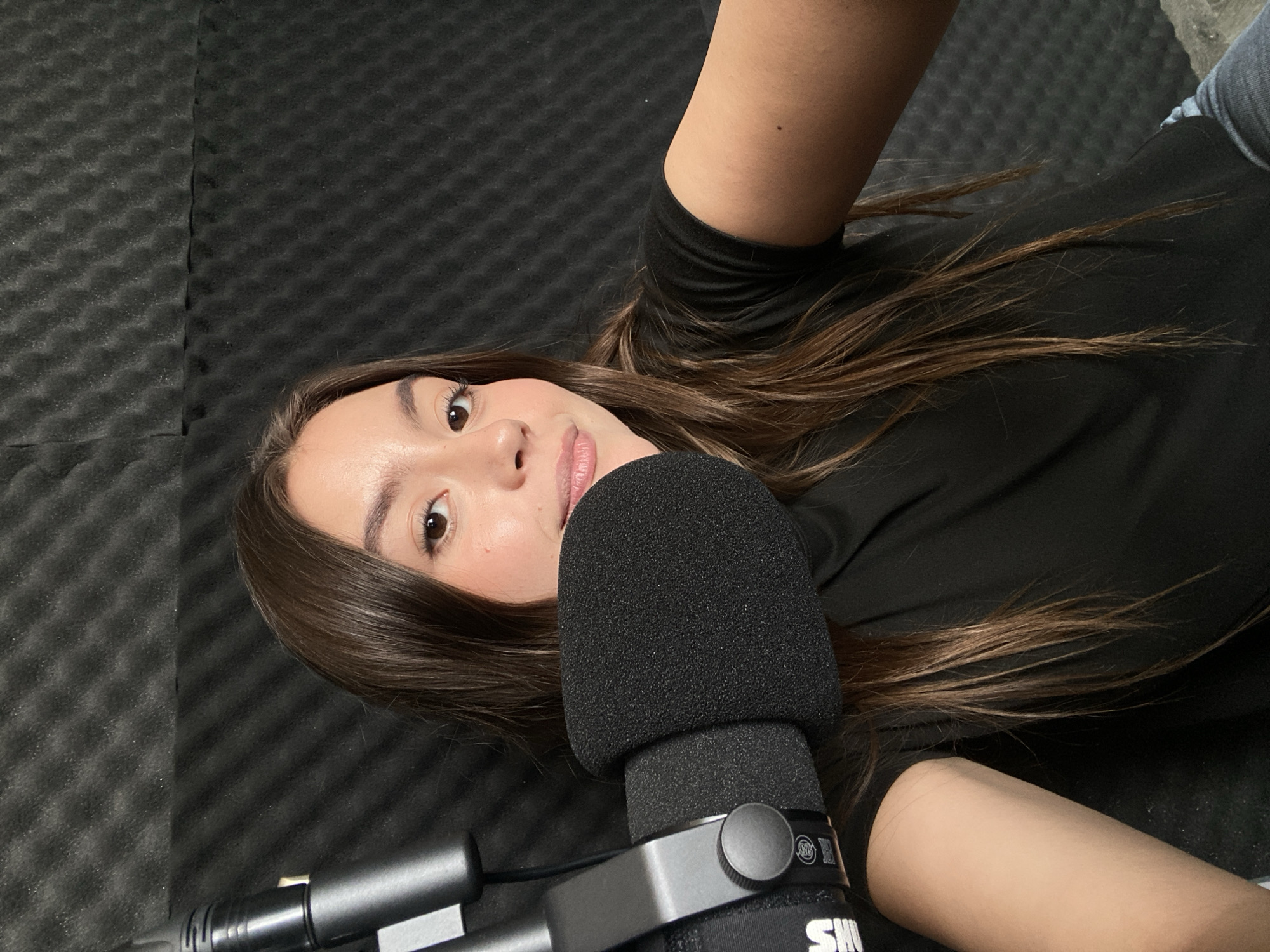 A young woman lies prone on a black sound-dampening foam surface, positioned in front of a Shure microphone with a foam windscreen. She wears a long-sleeved black shirt and appears to be in an audio recording studio. The city of Unknown, Unknown cannot be identified from the image.