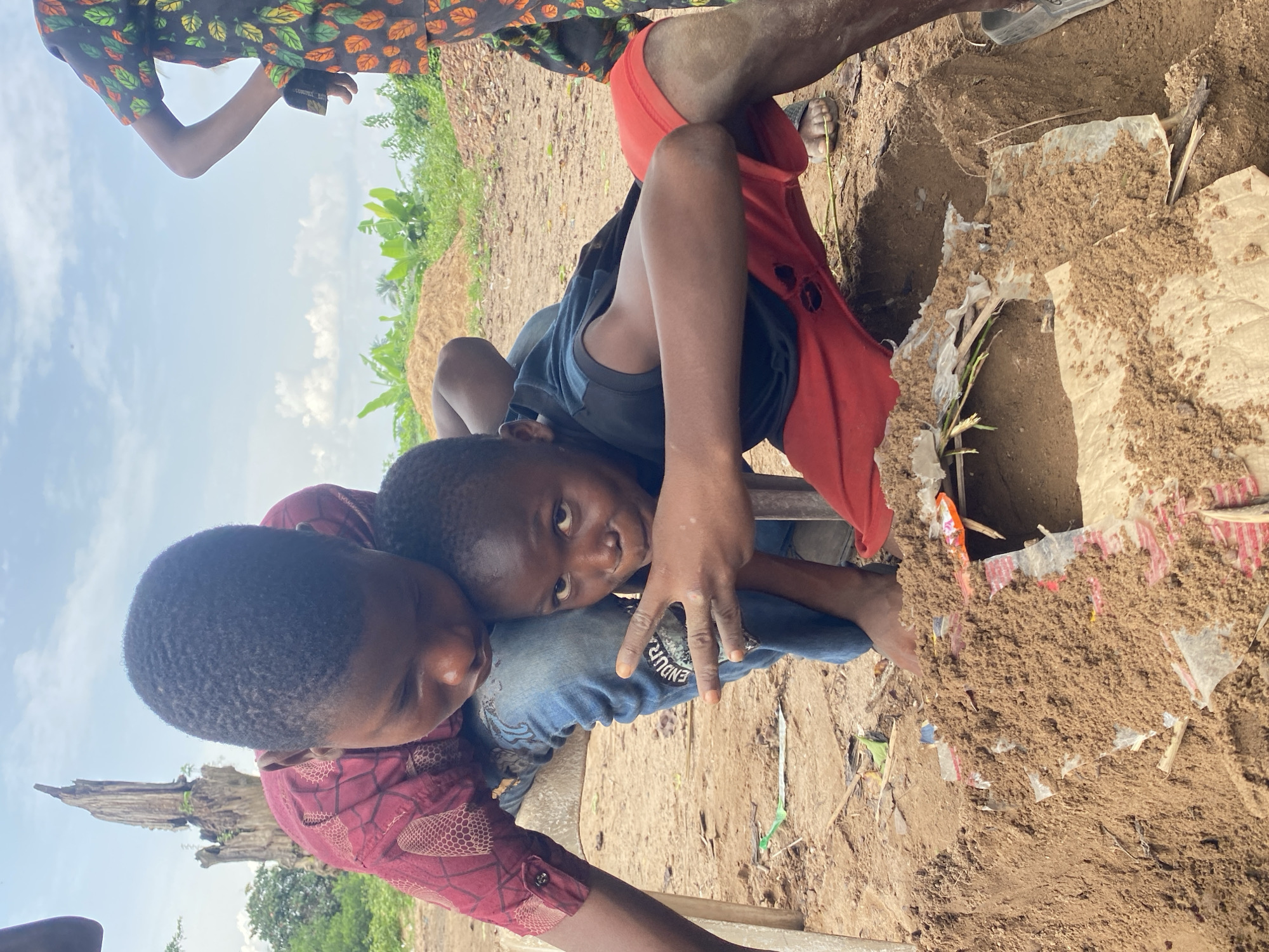 Two children are interacting near a dirt hole in Igbara, Nigeria. One child wears a maroon geometric patterned shirt and rests their head on the other child's knee. The other child wears a dark blue shirt and blue jeans with text on the leg. The hole is surrounded by loose dirt and scraps of white plastic and paper. Other people are partially visible in the image, including one person wearing a leaf-patterned shirt. A green field and blue sky with white clouds are visible in the background.