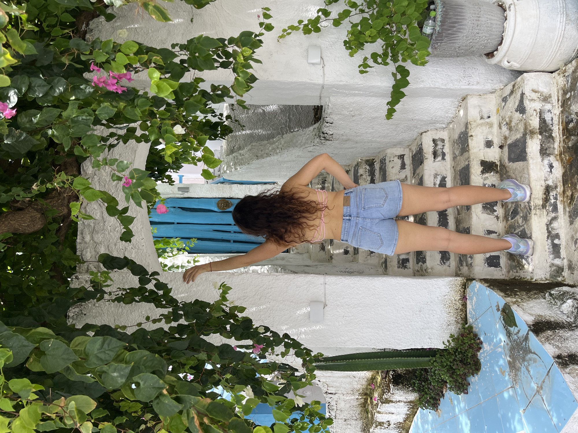 A woman wearing a pink bikini top and denim shorts stands with her back to the camera, facing a bright blue door set within a white arched entryway. She is reaching her left arm up towards a branch of green leaves and pink flowers that drapes over the archway. She is on a set of stone stairs that lead up to the door, with a blue tiled area to her left and a large potted plant to her right. The scene is in a visually rich, outdoor setting, possibly a courtyard or garden, characterized by lush greenery, whitewashed architecture, and vibrant floral accents.