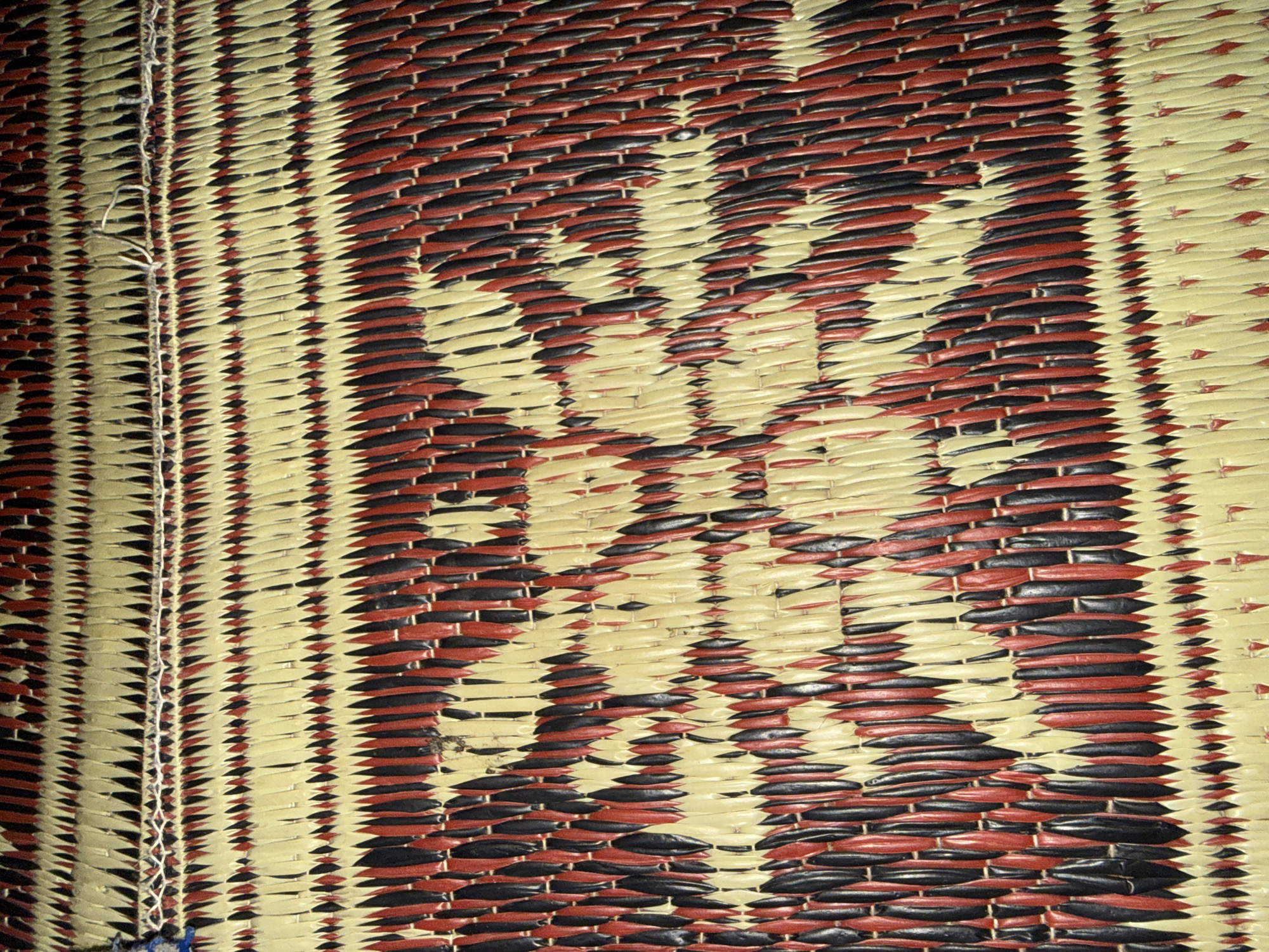The image shows a close-up view of a woven mat with a repeating pattern of red, black, and light tan colors. The weave is tight and creates a textured surface. There is a horizontal seam of white thread visible across the upper portion of the mat, suggesting it may be two sections joined together or an edge detail. The pattern resembles abstract floral or star-like motifs. The lighting is somewhat dim, casting subtle shadows that highlight the texture of the weave. The mat is likely made from natural materials like straw or reeds. The image is taken in Slahung, Indonesia, and depicts a common handicraft or decorative item found in the region.