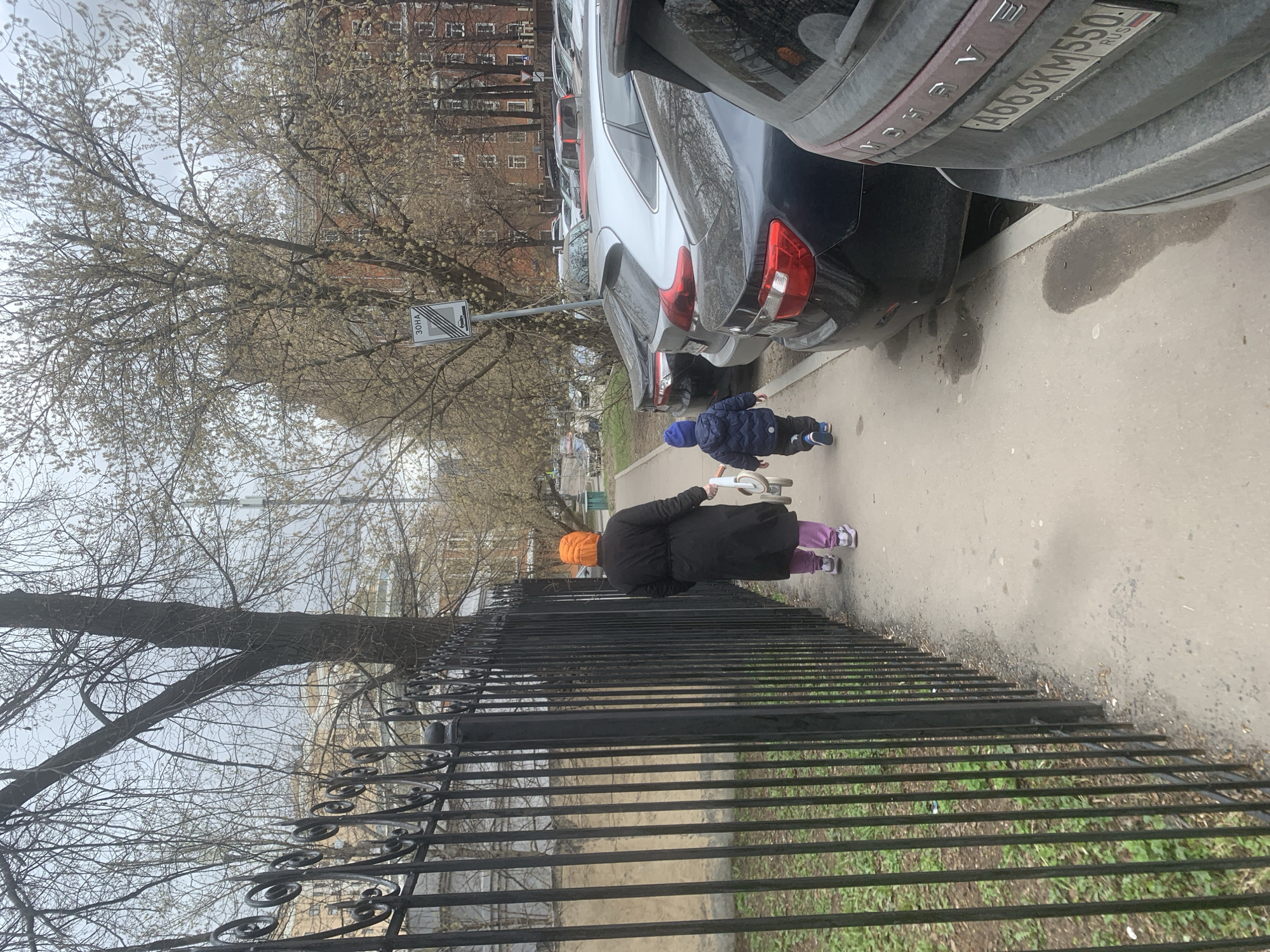 A street scene in Moscow, Russia, shows an older woman in a black coat and orange hat walking along a sidewalk with a young child in a blue coat and hat. The child is slightly ahead of her. The woman is pushing a white toy. A black wrought iron fence runs along the left side of the frame, separating the sidewalk from a grassy area. Parked cars line the right side of the sidewalk. The license plate A663KM550 RUS is visible on one of the cars. Bare trees are visible in the background, and a building is in the distance. A street sign is also present. The sky is overcast.