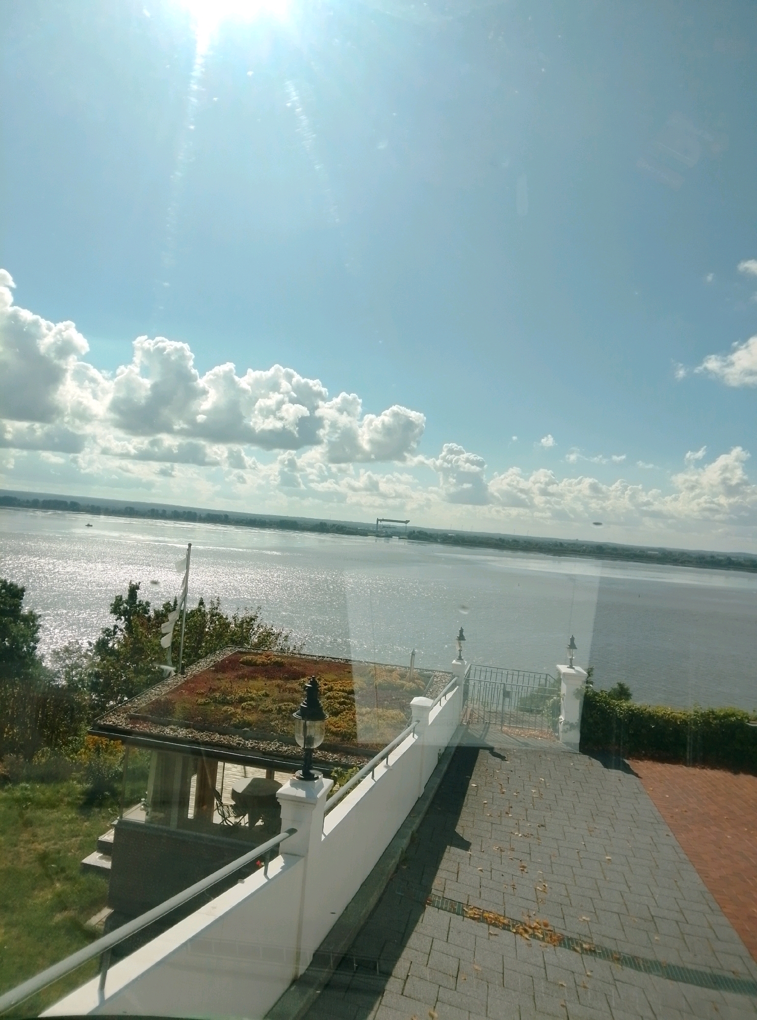 The image captures a bright, sunny day overlooking a wide body of water, likely the Elbe River, in Hamburg, Germany. The sun is high in the sky, casting a strong glare and creating bright reflections on the water's surface. Cumulus clouds are scattered across the light blue sky.
In the foreground, there is a paved walkway with fallen autumn leaves. A white railing separates the walkway from a lower grassy area and a small building with a green, vegetated roof. A black lamppost stands on a white pedestal next to the railing. Further back, on the grassy area, a small structure with glass walls and a peaked, vegetated roof is visible. A flagpole with white flags stands behind this structure.
Across the water, a distant shoreline with trees and structures, including what appears to be a large industrial crane or bridge, can be seen. A few small boats are visible on the water. The overall impression is a serene, waterside view on a clear, autumn day.