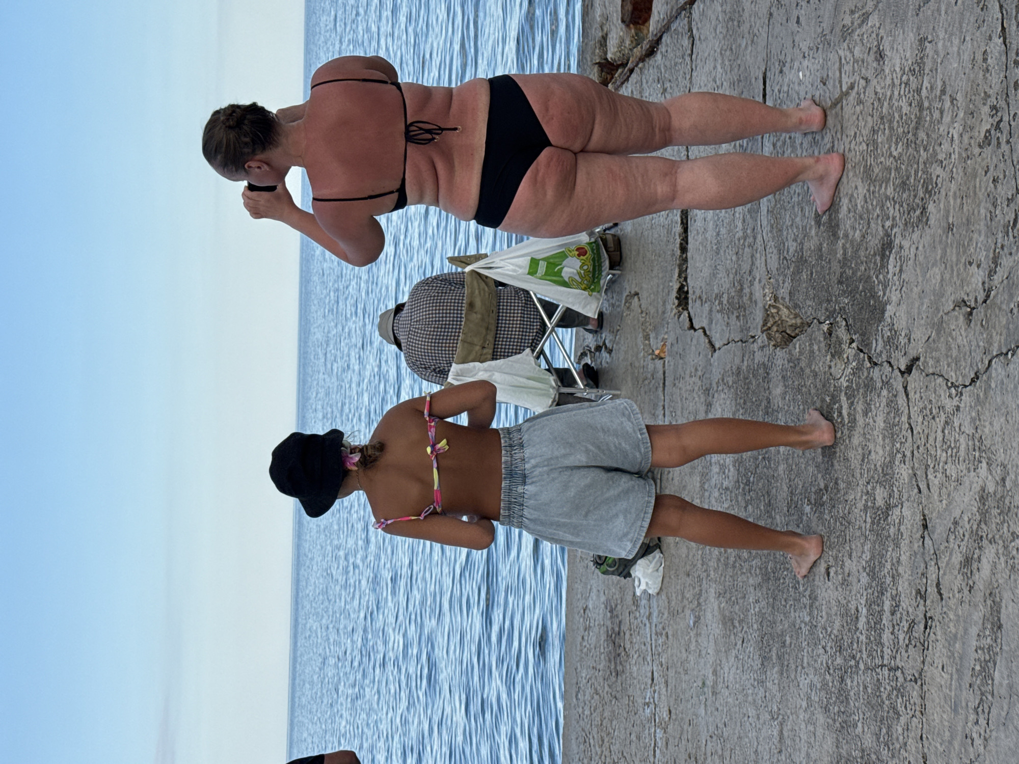Two individuals are on a concrete surface next to a body of water. The person closer to the viewer is a young female wearing a black hat, a colorful bikini top, and light gray shorts. She is standing with her legs slightly apart, facing away from the camera. To her left, a folded white cloth is visible. Behind her, a person wearing a plaid shirt and a hat is seated in a folding chair. A white plastic bag with green lettering is hanging on the back of the chair. Further back, a woman in a black bikini is standing and holding a dark object to her face. The scene appears to be outdoors, with a clear blue sky and calm water. The concrete surface is weathered and cracked. The lighting suggests it is daytime, possibly sunny. The overall mood seems relaxed and casual, typical of a waterside location. No discernible text is visible other than on the plastic bag, which is partially obscured. The location is identified as Partenit, Ukraine.