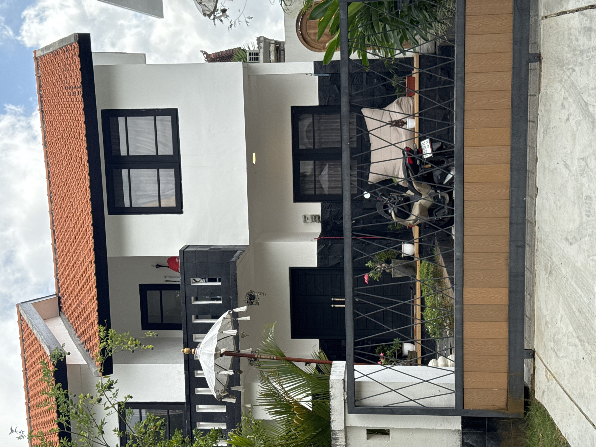 The image shows a two-story white house with an orange tiled roof in Kuta Selatan, Indonesia. The house has black window frames and black decorative elements. A black metal gate with a wooden inlay separates the property from the street. Inside the gate, a motorcycle is parked next to a large beige cushion on a bench. Several potted plants are visible on a shelf behind the bench. A decorative white umbrella on a stand is positioned in front of the house, to the left of the main entrance. The sky is cloudy.
