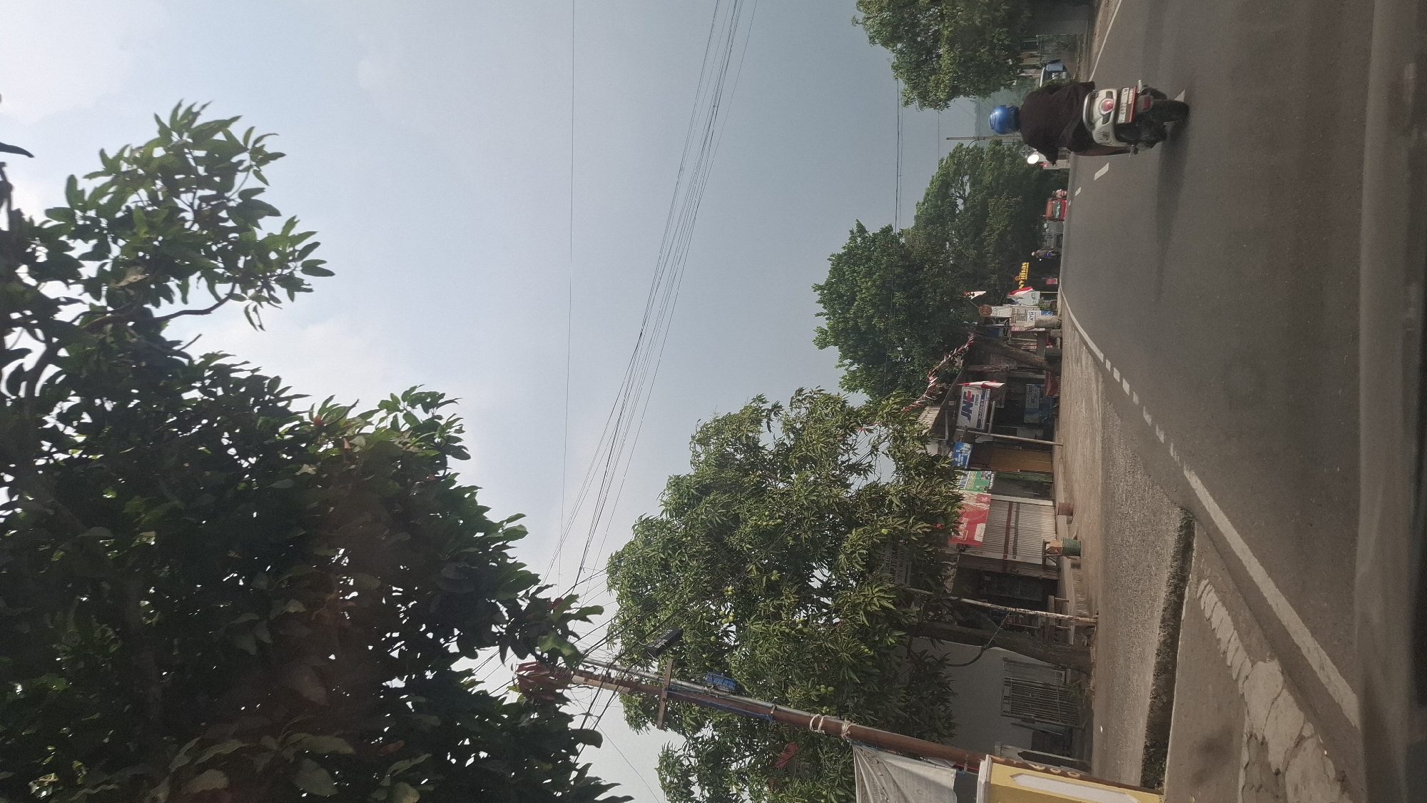 A person wearing a blue helmet and a dark jacket rides a white scooter down a paved road in Susukan, Indonesia. The scooter has a license plate that reads "AD 3338 GBN". The road is flanked by small shops and trees on the left side and more trees on the right. Power lines crisscross the sky, which is overcast. The scene appears to be a typical street in a town or village setting, with a blend of residential and commercial structures.