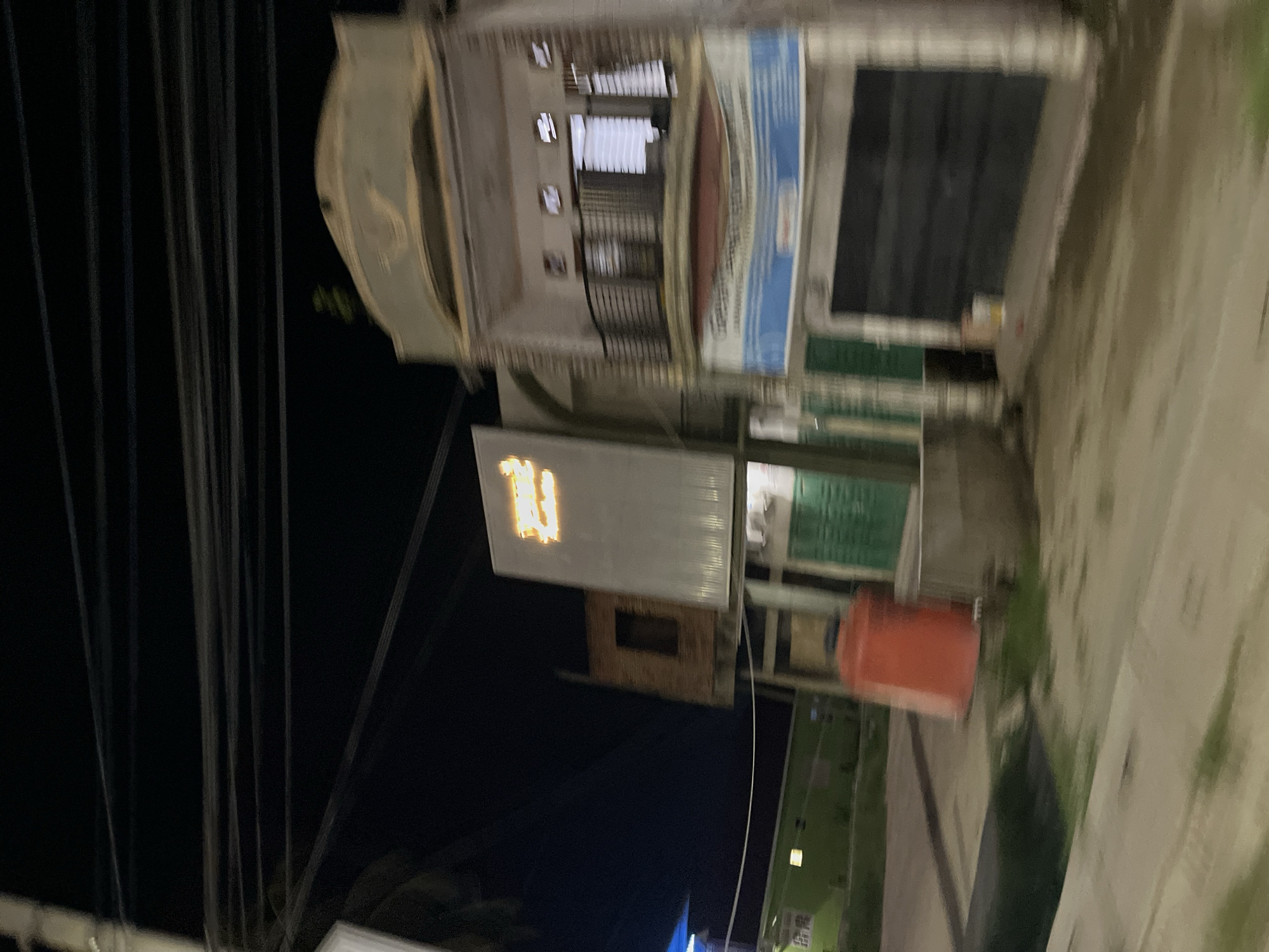 The image depicts a blurry nighttime street scene in Bundar, Indonesia. In the foreground, a concrete sidewalk is visible, with a dark, wet area suggesting recent rain or a ditch. To the left, a green building with illuminated windows can be seen. Further back, an orange water tank stands next to a building under construction.
The main focus of the image is a multi-story building on the right. The lower level appears to be a shop with a large, dark shuttered entrance. Above this, a section with a blue and white banner is visible. The second floor has a balcony with a dark railing and several windows. The top of the building features an arched architectural element.
Above the buildings, a dense network of power lines stretches across the dark sky. A brightly lit sign with orange lettering is affixed to a facade on the left side of the street, partially obscuring the building underneath. The overall impression is of a modest urban environment at night, characterized by a mix of commercial and residential structures.