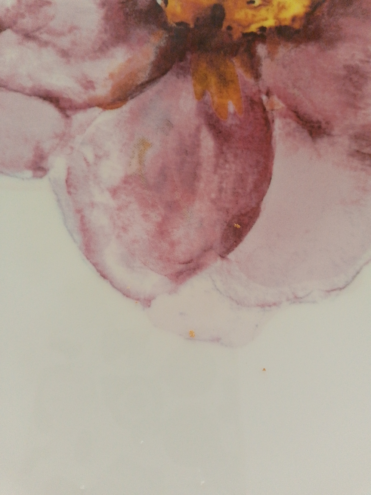 A close-up, overhead shot captures a watercolor painting of a flower. The flower's petals, in soft shades of pink and white, unfurl towards the center, where warm yellow and brown tones suggest the stamen and pistil. The painting has a delicate, ethereal quality, with blurred edges and subtle color variations creating a sense of depth and movement. A few tiny flecks of gold leaf are scattered across the canvas, adding a touch of sparkle to the artwork.