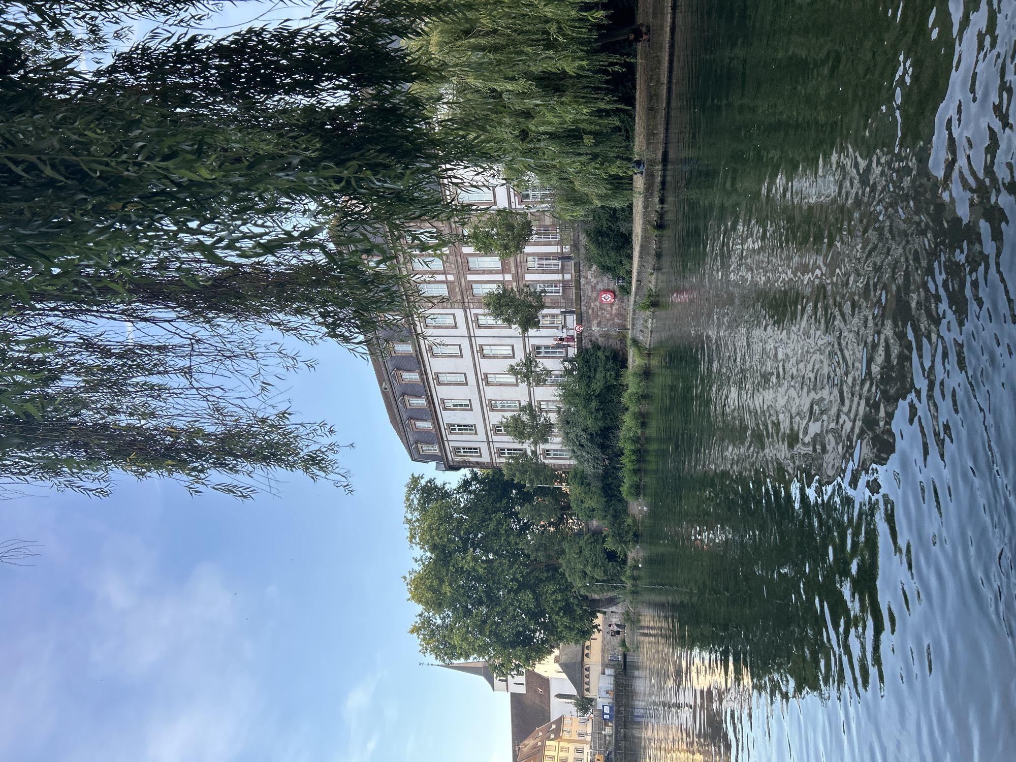 The image depicts a scenic view of the Ill River in Strasbourg, France, with the Petite France district visible in the background. A large, multi-story building with a white facade and brown trim stands prominently on the right bank of the river, characterized by numerous windows and a mansard roof. To the left of this building, a church steeple with a cross pierces the sky, suggesting its religious significance.
The river itself is calm, reflecting the surrounding architecture and lush greenery. Willow trees, with their distinctive drooping branches, frame the scene, their leaves creating a dappled effect against the blue sky. The riverbank is lined with vegetation and a paved walkway, where several individuals are seated or strolling. One person is visible near the building, appearing to be walking along the river, while others are seated on the bank, enjoying the serene atmosphere. A red life preserver is affixed to the wall near the river, a common safety feature along waterways. The overall ambiance is peaceful and picturesque, capturing a moment of tranquility in this historic European city.