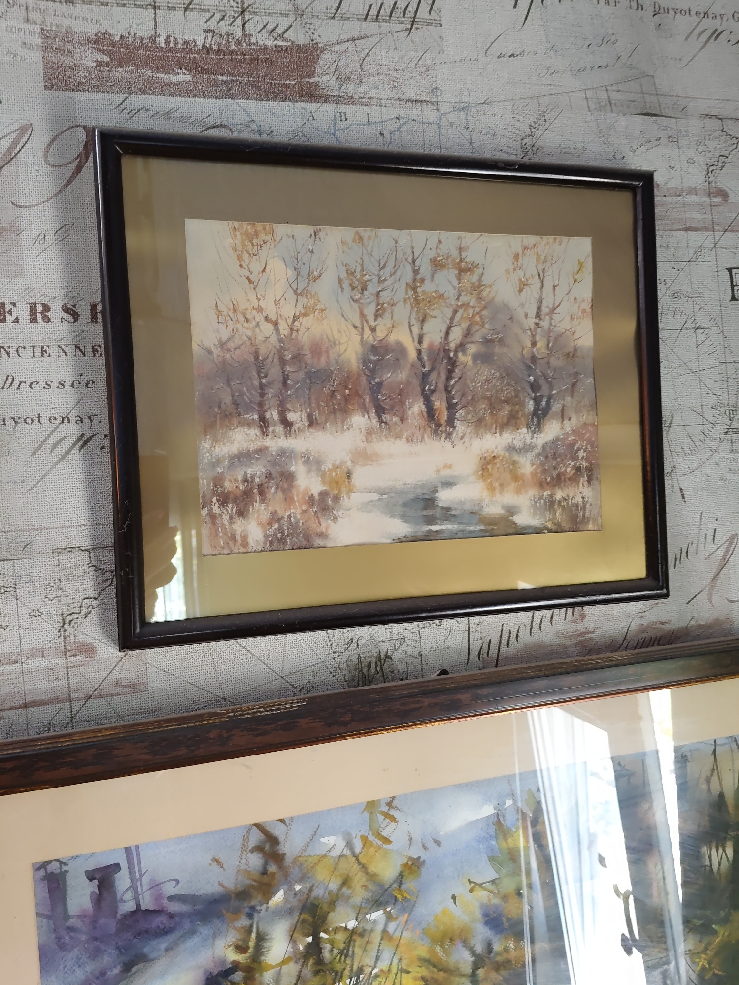 The image shows two framed artworks hanging on a wall with a patterned wallpaper. The upper artwork is a watercolor painting of a winter landscape with bare trees and a partially frozen stream. The lower artwork, only partially visible, appears to be another watercolor painting of a landscape with trees and foliage. The wallpaper has a faded design resembling old maps and script. The lighting suggests an indoor setting, possibly a home or gallery. There is no discernible information in the image that would indicate its location in the city of Rostov-on-Don, Russia.