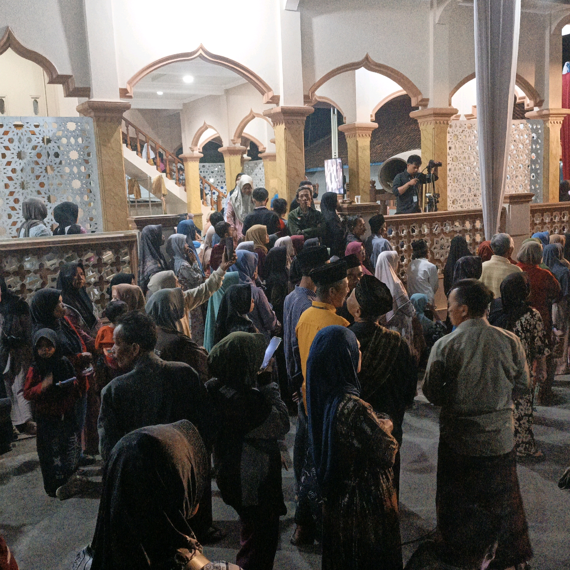 In the city of Kalitorong, Indonesia, a gathering of people is observed within an indoor space that appears to be a mosque or a similar community building. The architecture features prominent arched openings and pillars with decorative elements. A staircase is visible in the background, suggesting multiple levels within the structure.
The scene is populated by a diverse group of individuals, many of whom are wearing traditional Islamic attire, including hijabs and modest clothing. Some people are standing and conversing, while others are seated or in motion. In the foreground, a person is partially visible from behind, facing away from the camera. To their left, another individual is visible from the side, wearing a headscarf.
Further into the scene, a man is seen operating a video camera mounted on a tripod, indicating that an event is being filmed. Several groups of people are clustered together, suggesting social interaction or participation in a shared activity. The lighting is artificial, creating a warm ambiance. The ground appears to be paved, and decorative screens or partitions are visible along some edges of the space. The overall atmosphere suggests a community event or religious gathering.