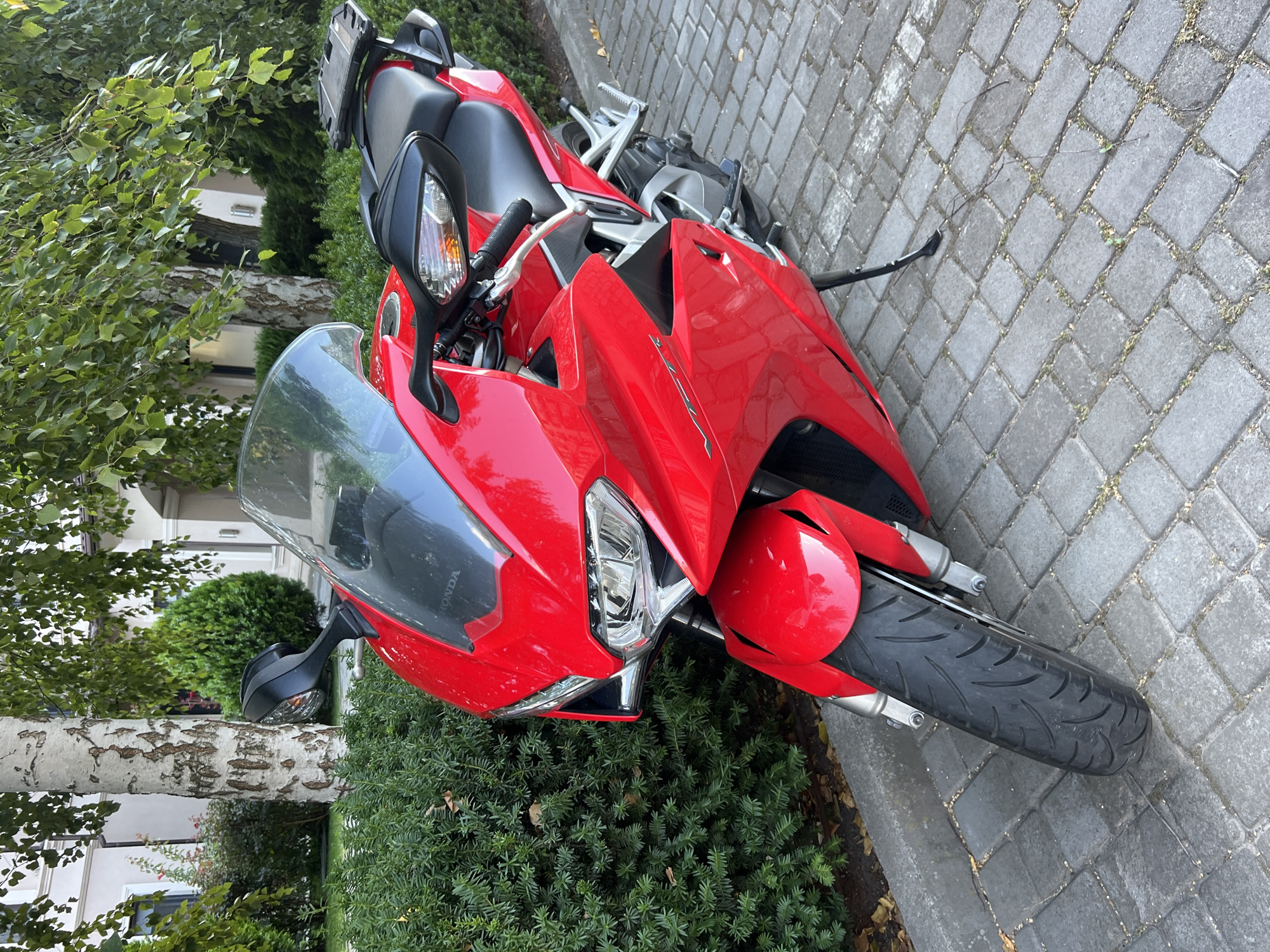 A red Honda motorcycle is parked on a cobblestone pathway in front of a building with trees and bushes. The motorcycle is positioned at an angle, with its front wheel closer to the camera. It has a black seat, a silver frame, and a transparent windshield. The background shows lush green foliage and a building with light-colored walls and white columns. The environment appears to be a well-maintained outdoor space, possibly a residential or commercial area in Fontanka, Ukraine. No people are visible in the image.