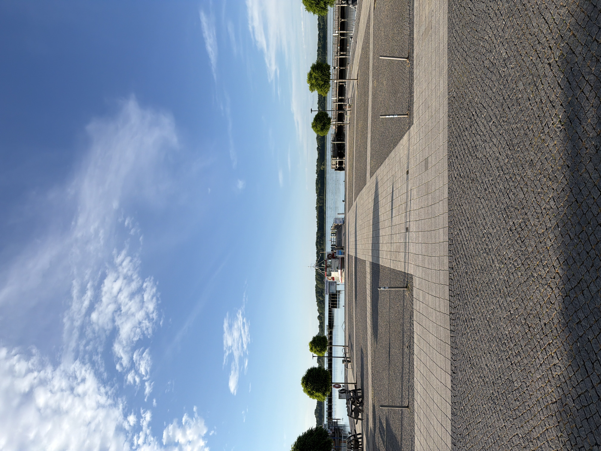 The image depicts a waterfront promenade in Ribnitz-Damgarten, Germany. The scene is oriented vertically, with the sky occupying the left portion and the promenade and water on the right. The sky is a vibrant blue, with scattered white clouds, suggesting fair weather and daytime. The promenade is paved with two different materials: a section of light-colored square tiles and a section of dark cobblestones. Several trees, pruned into rounded shapes, are planted along the promenade, with benches placed between them.
In the background, a body of water is visible, with a tree-lined shore on the opposite side. A white boat with a red stripe and the text "Schiffsausflugshafen" (meaning "Excursion Boat Harbor" in German) is docked near the promenade. The presence of the boat and the harbor text clearly indicates a location related to water transportation and tourism. The time of day appears to be late morning or early afternoon, given the angle of the shadows cast by the trees and posts on the promenade. The overall atmosphere is peaceful and suggests a pleasant day for a stroll or relaxation by the water. There are no people visible in the image.
