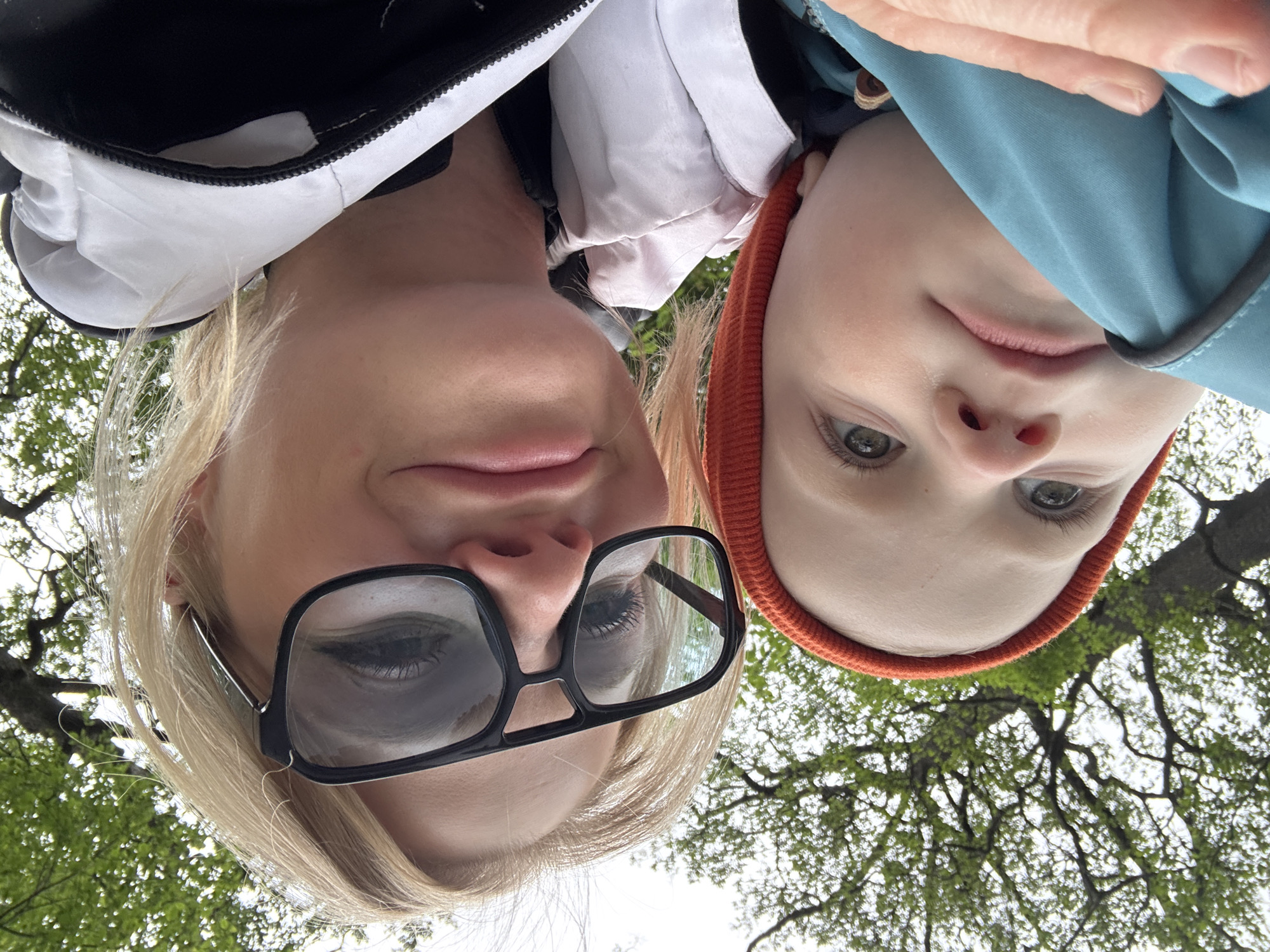 A selfie taken from a low angle, looking up at two individuals, likely a woman and a child, set against a backdrop of trees. The woman, on the left, has blond hair and is wearing glasses. The child, on the right, is wearing a hat and jacket. The trees in the background suggest an outdoor setting, potentially a park or green space. Although I don't know the exact location, there are many of the parks in Saint Petersburg, Russia, that could fit this description.