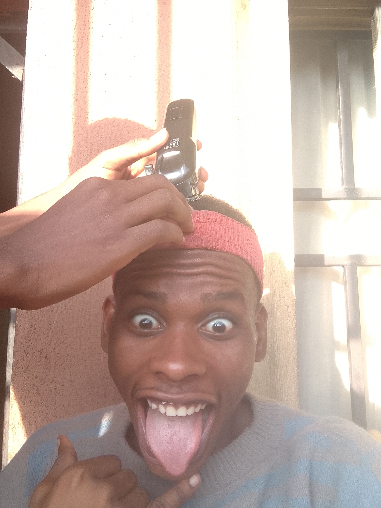 A man is receiving a haircut outdoors in the city of Unknown, Unknown. The man has dark skin, wide eyes, an open mouth with his tongue sticking out, and is wearing a red headband. He is wearing a grey sweater. A hand holding a black electric hair clipper is above his head, seemingly cutting his hair. The background consists of a beige-colored wall and a window. The scene is illuminated by bright sunlight, causing shadows on the wall.