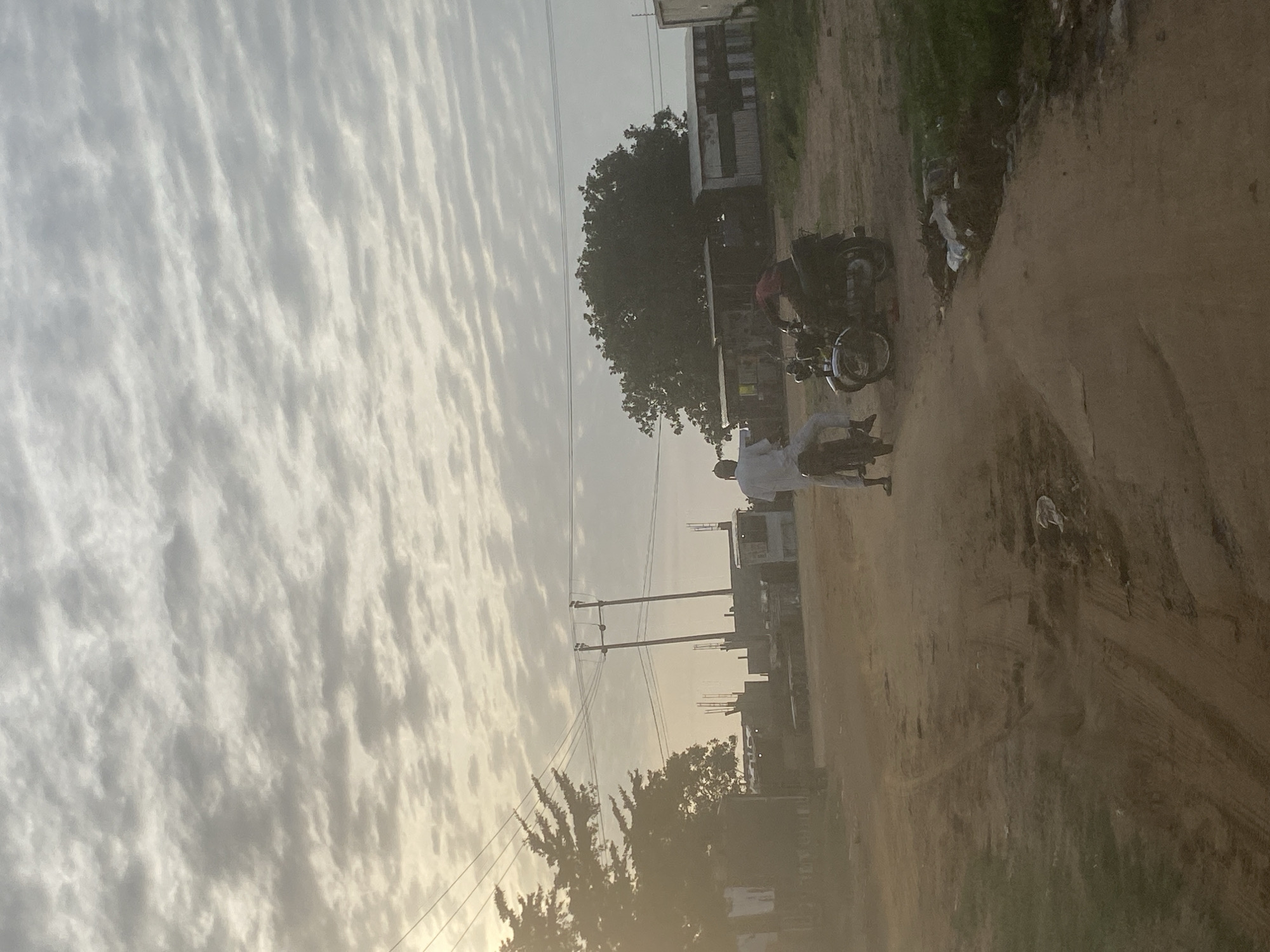 The image depicts a dirt road in Lugbe, Nigeria. Overcast skies fill the background. A person in a white shirt is on a motorbike. Another person in a red shirt stands near a second motorbike. Trees and buildings are visible on either side of the road, alongside power lines. Debris litters the roadside.