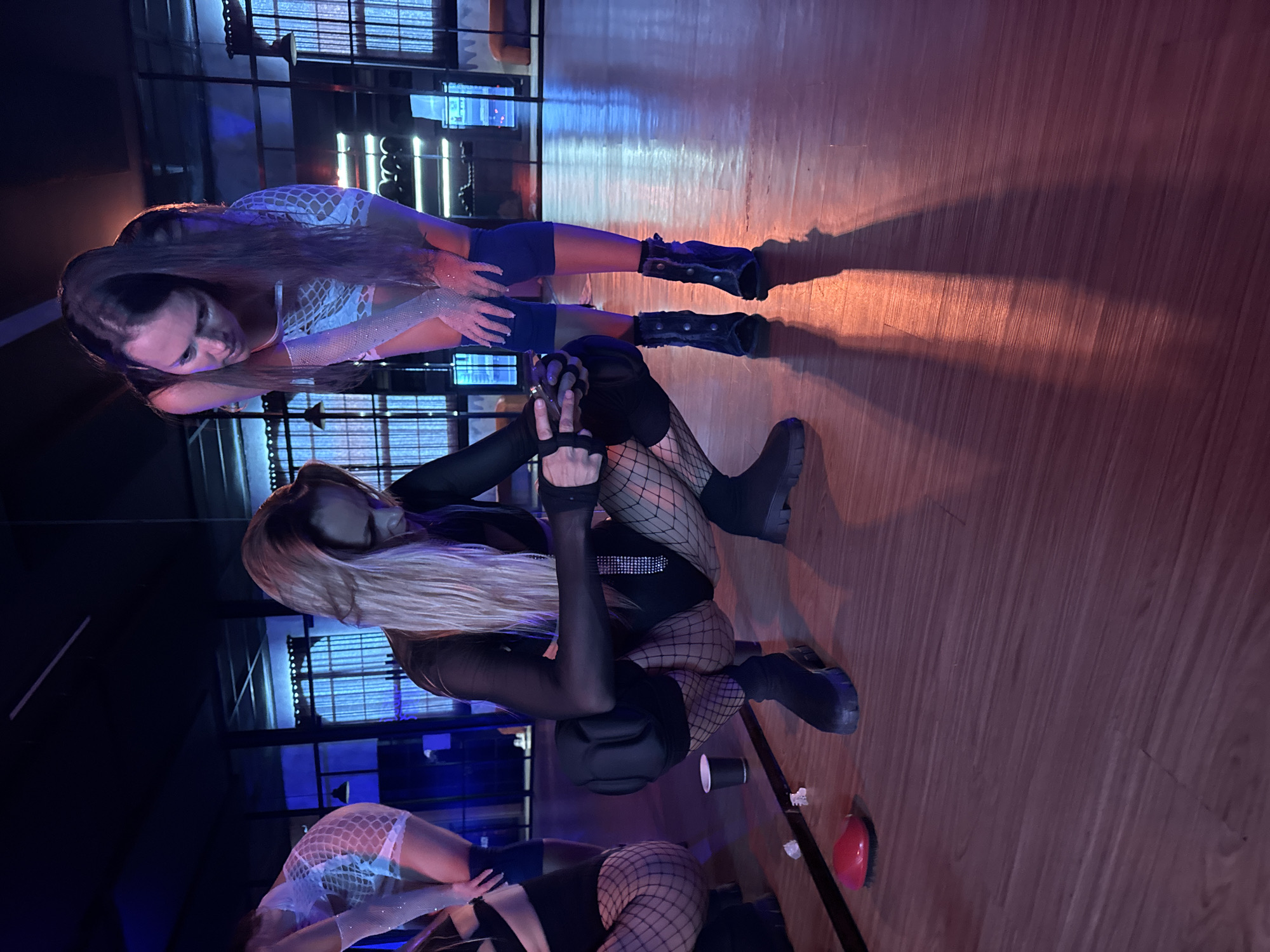 The image depicts two individuals in what appears to be a dance or performance setting. One individual, with long blonde hair, is kneeling and wearing a black outfit with fishnet stockings, fingerless gloves, and platform boots. They have knee pads on. The other individual has long dark hair and is wearing a short dress with what appears to be a fishnet overlay, short shorts, arm warmers, and boots. They are positioned near a mirrored wall. The setting appears to be indoors, with wooden flooring, and is lit with blue and orange lighting. There are cups and other small items scattered on the floor. The location is suspected to be in the city of North Kuta, Indonesia.