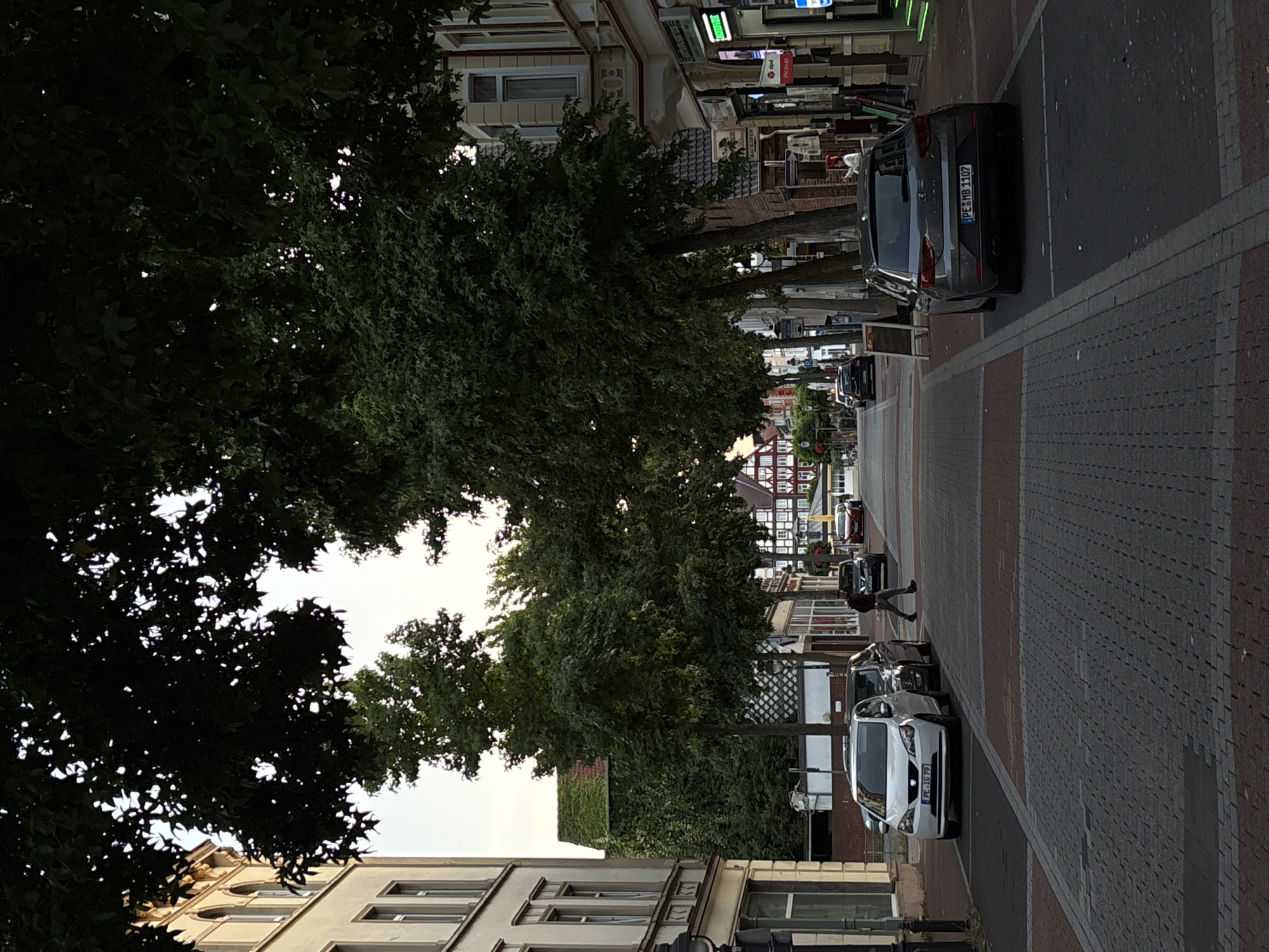 In the city of Unknown, Unknown, a street scene unfolds beneath the canopy of leafy trees, casting dappled shadows on the paved road. On the left, a light-colored, four-door car is parked along the sidewalk, its license plate reading "PE JG 79". To its right, a darker car is also parked. A pedestrian, dressed in dark clothing, walks across the street from left to right. Further down the road, a dark-colored, five-door hatchback is parked on the right side, its license plate displaying "PE MB 1107". In the distance, more cars are visible on the street, and buildings line both sides of the thoroughfare, some featuring traditional architectural styles with visible timber framing. The sky above is a pale, overcast hue.