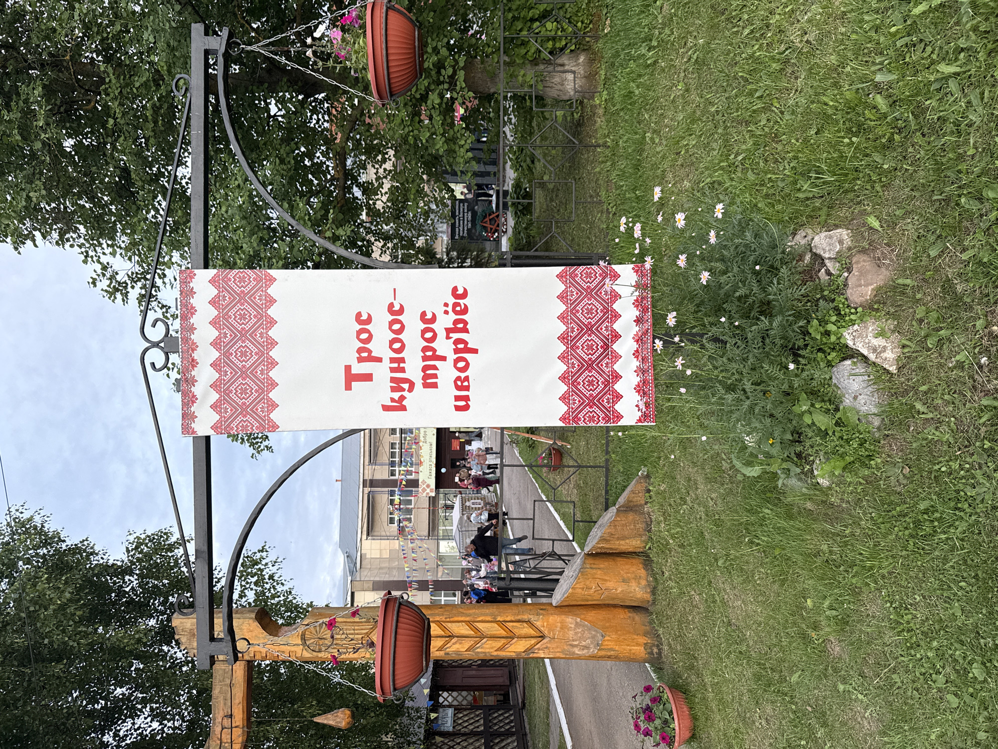 The image shows a banner with red and white ethnic patterns and text in Russian hanging from a metal archway supported by wooden posts. The banner reads "Трос купос-трос иорвёс". In the background, there is a building decorated with colorful flags. A group of people are gathered in front of the building. There are potted plants with flowers hanging from the archway and on the ground. The scene is set outdoors with a grassy area and trees. The provided coordinates place the image in the village of Kukuy, Russia.