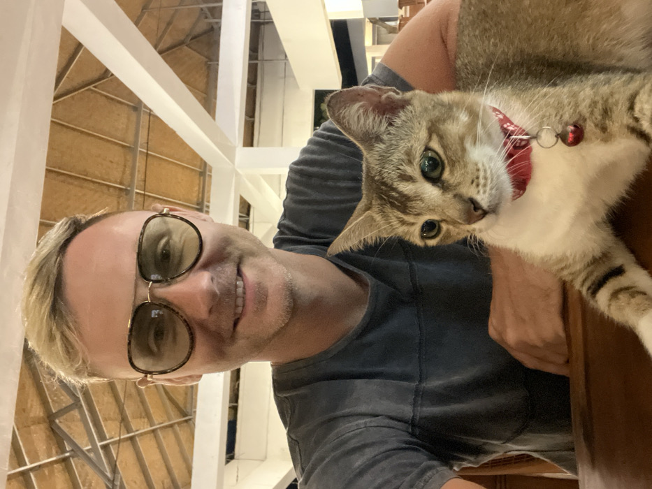 An adult male and a tabby cat are depicted in a close-up image taken indoors or within a semi-open structure in Uluwatu, Indonesia. The male, positioned on the left, has short blond hair, is wearing aviator-style sunglasses with dark frames and light-tinted lenses, and a grey t-shirt with horizontal darker stripes. He is smiling directly at the camera. A light brown and white tabby cat, wearing a red collar with a small red bell, sits on a wooden table in front of the male, looking slightly off-camera to the right. The background reveals white structural beams supporting a thatched or woven natural material ceiling. Out-of-focus wooden furniture is visible in the distant background.