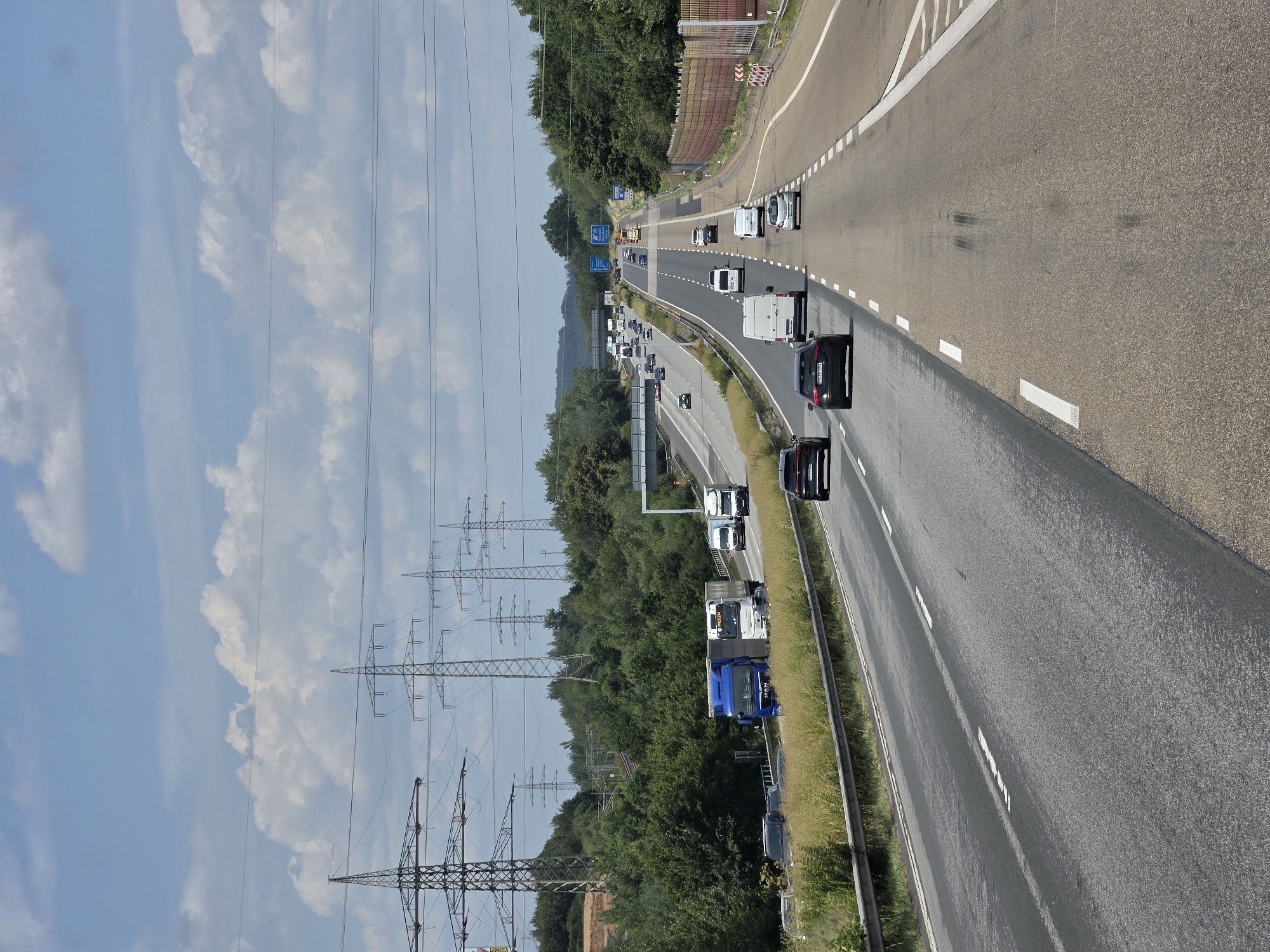 The image depicts a sunny day on a multi-lane highway with traffic. In the foreground, the road surface is visible, with lane markings. Several cars and vans are driving on the highway. In the middle ground, more vehicles are visible, including trucks. In the background, a dense line of trees borders the highway, and tall electrical pylons are prominent against a blue sky with white clouds. The scene is likely on a highway near Ebernhahn, Germany, given the presence of German license plates on some vehicles and the general landscape, which is consistent with the region. There are no people clearly visible in the image. The overall impression is one of typical highway traffic flow in a rural or semi-rural setting.