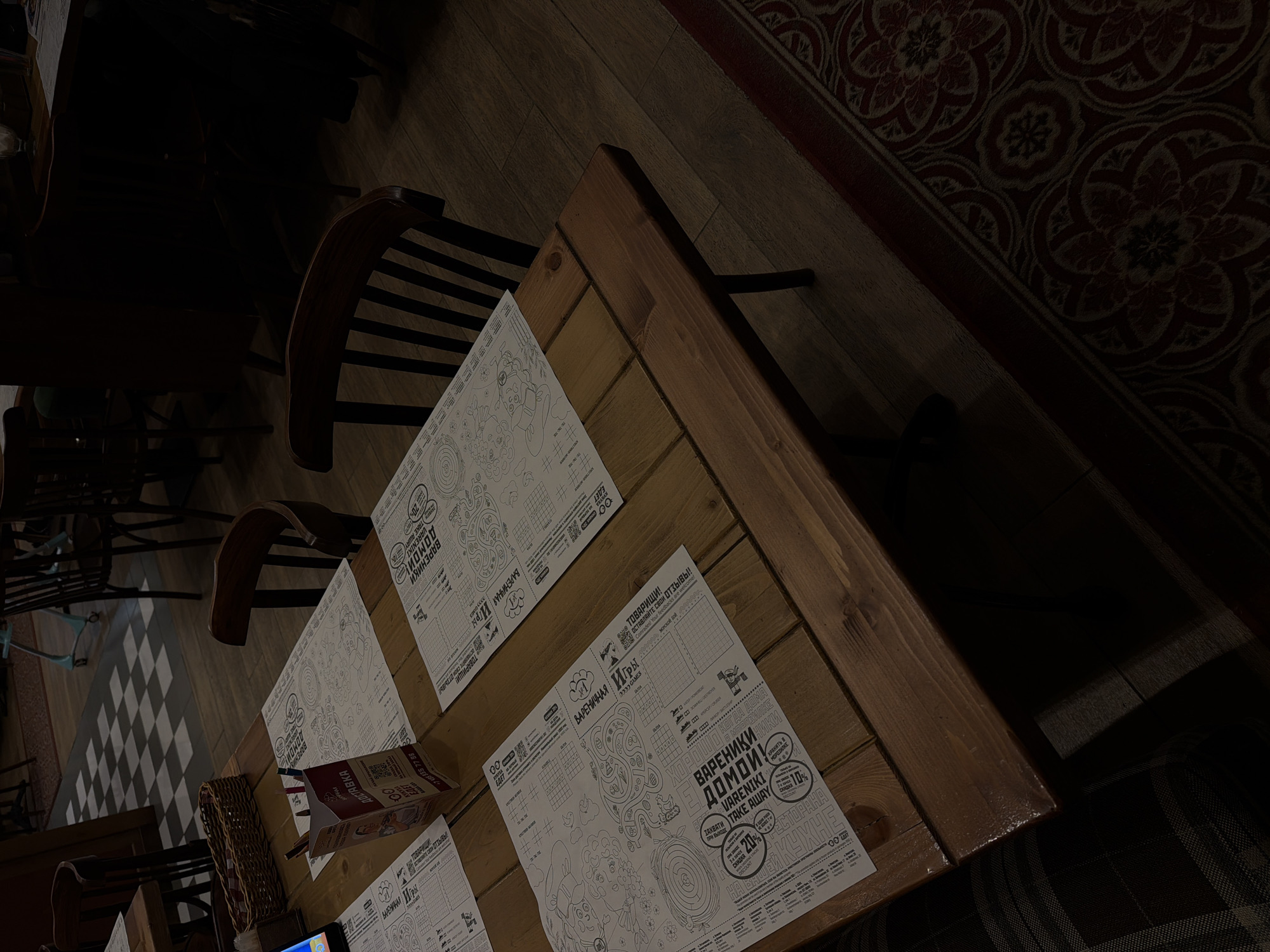 The image presents a dimly lit interior of what appears to be a Russian restaurant or cafe, likely a "Varenychna" (dumpling house), located in Moscow, Russia. The scene is captured from an overhead angle, centering on a robust wooden table where no people are present. Several dark wooden chairs are visible, including one partially in the foreground and another fully in the midground, both featuring slatted backs.
The table is set with multiple paper placemats, designed with intricate line art for coloring, grid-based games, and promotional text, all primarily in Cyrillic. A wicker basket rests on the table, possibly holding condiments or cutlery. The floor under the table consists of dark wooden planks. To the right, a dark red rug with a traditional floral pattern covers the floor, while in the background on the left, a section of a black and white checkered tiled floor is visible, along with glimpses of other tables and chairs, including a light blue chair base. The low lighting suggests an evening setting or a cozy, atmospheric interior.
Visible text on the placemats includes phrases such as "ВАШЕЙ КУХНЕ" (YOUR KITCHEN), "ВАЛЕНКИ ДОМОЙ" (VARENIKI HOME), indicating take-away dumplings with instructions to "ХРАНИТЬ В МОРОЗИЛКЕ" (STORE IN FREEZER). Promotions mention "TAKE AWAY" with discounts of "10%" and "20%". The placemats are branded "ВАРЕНИЧНАЯ ИГРЫ" (VARENYCHNA GAMES) and feature calls to action like "ТОВАРИЩИ! ОСТАВЛЯЙТЕ СВОИ ОТЗЫВЫ!" (COMRADES! LEAVE YOUR REVIEWS!). QR codes are also printed on the placemats.