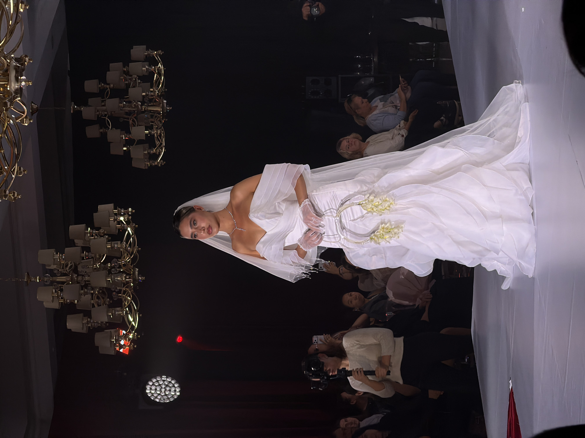 A fashion model walks a runway while wearing a white wedding dress. The model is wearing a white veil and white gloves. She is also wearing a diamond necklace. She is surrounded by spectators. Two golden chandeliers are visible. A white wall is visible. There are several people taking pictures of the model. The location is in the city of Unknown, Unknown.