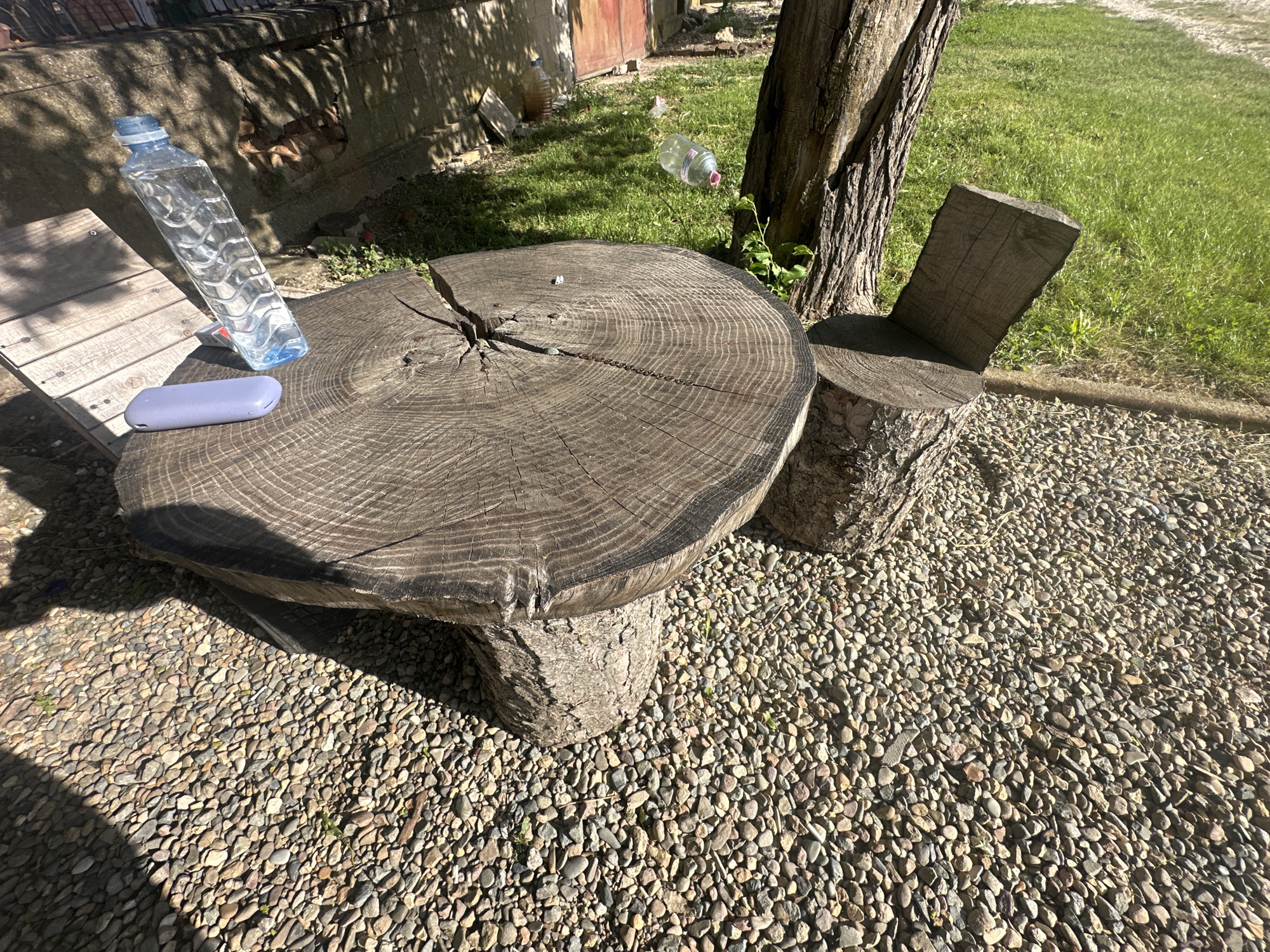 The image shows an outdoor scene, likely a yard or garden, situated in Sirakovo, Bulgaria. A rustic table is the central focus, crafted from a large, circular tree stump with visible growth rings and cracks. The table is supported by a smaller stump base. Next to the table, a rudimentary chair is constructed from tree stumps, including a backrest section. On the table, a clear plastic water bottle is present, along with a lavender-colored electronic device (possibly a phone or portable charger). The ground surrounding the table and chair is covered in small pebbles or gravel. Green grass is visible in the background near a tree trunk. An outdoor building can be seen in the distance. The scene is bathed in sunlight, creating shadows around the table and chair.