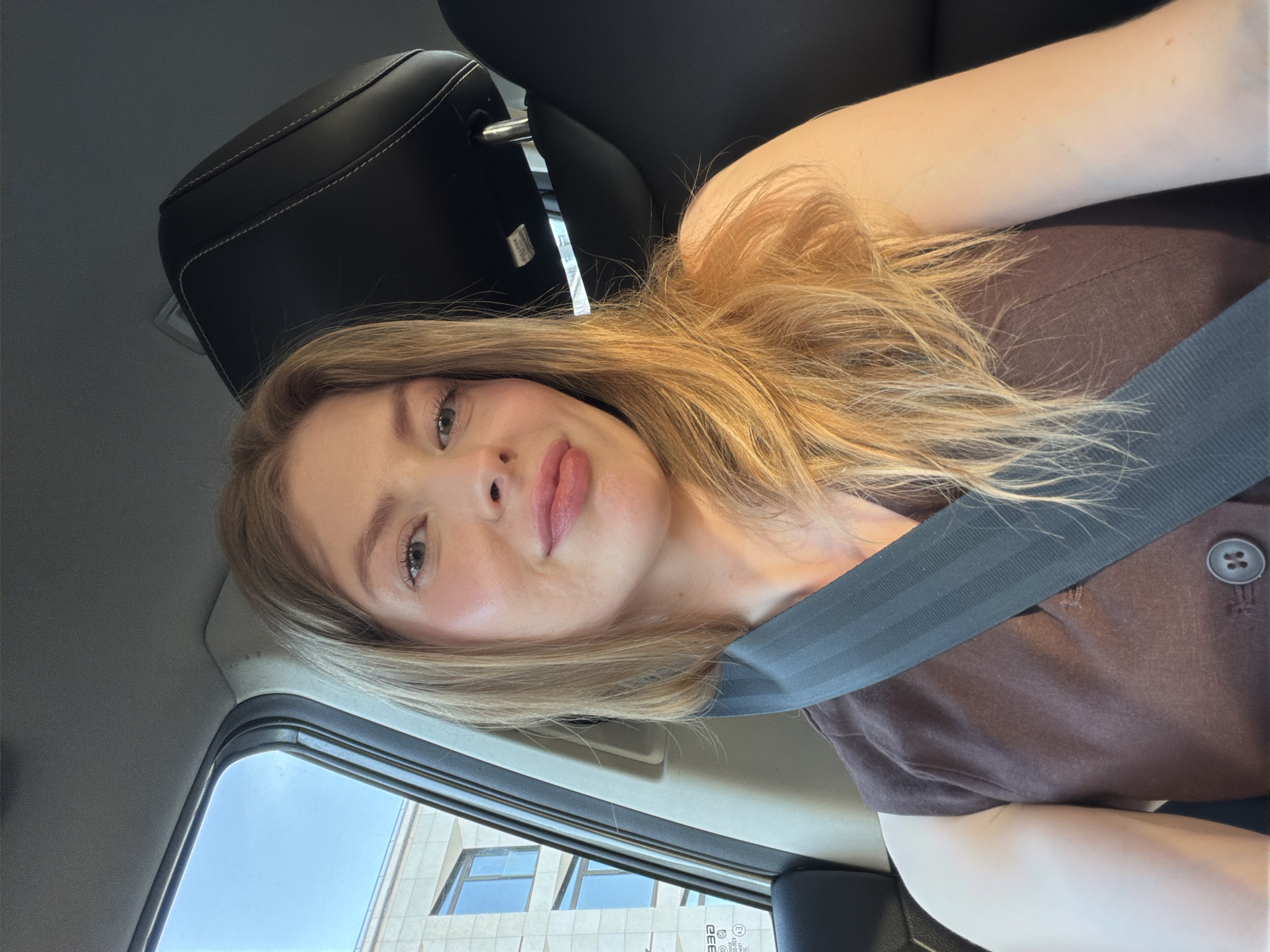 A young woman is captured in a self-portrait taken inside a car, positioned as a passenger. She has long, wavy light brown or blonde hair and fair skin, with light-colored eyes gazing directly at the camera. A soft, pleasant smile graces her lips. She is dressed in a simple brown short-sleeved top with a dark button on the placket, and a dark grey seatbelt crosses her chest. The car's interior is visible, featuring a black leather seat and headrest with distinct white stitching. Through the car window on her left, a clear blue sky and a modern urban building are discernible. The building displays light-colored stone or concrete blocks and dark-framed windows, characteristic of an urban environment. The bright, natural lighting indicates it is daytime with good weather. Faint text, including "CEE", "E3", and "332", is visible on a sticker or etching on the car window. This scene is set in Moscow, Russia.