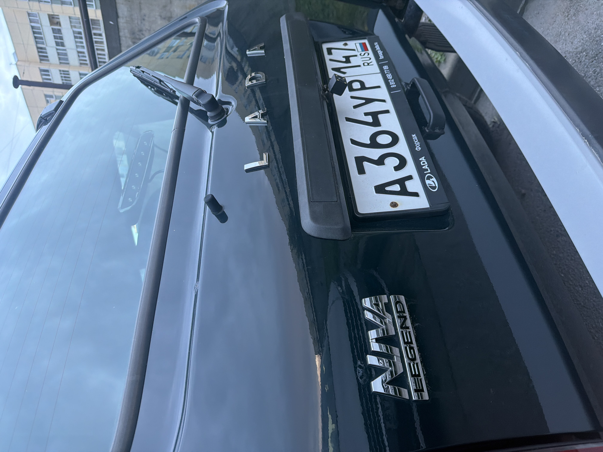 This media file shows the rear of a dark-colored LADA Niva Legend car. The focus is on the trunk area and the license plate. The license plate is white with black text and reads "A364YP147 RUS". Above the license plate, the LADA logo and name are visible in chrome lettering. Below the license plate, the "NIVA LEGEND" emblem is also visible in chrome. A small black camera is mounted just above the license plate. The car's rear windshield wiper is also visible. The scene appears to be outdoors, with a cloudy sky reflected in the car's surface. A building with multiple windows is partially visible in the background, suggesting an urban or suburban setting. There is no indication of people or a specific event taking place. The lighting suggests it is daytime, possibly overcast.