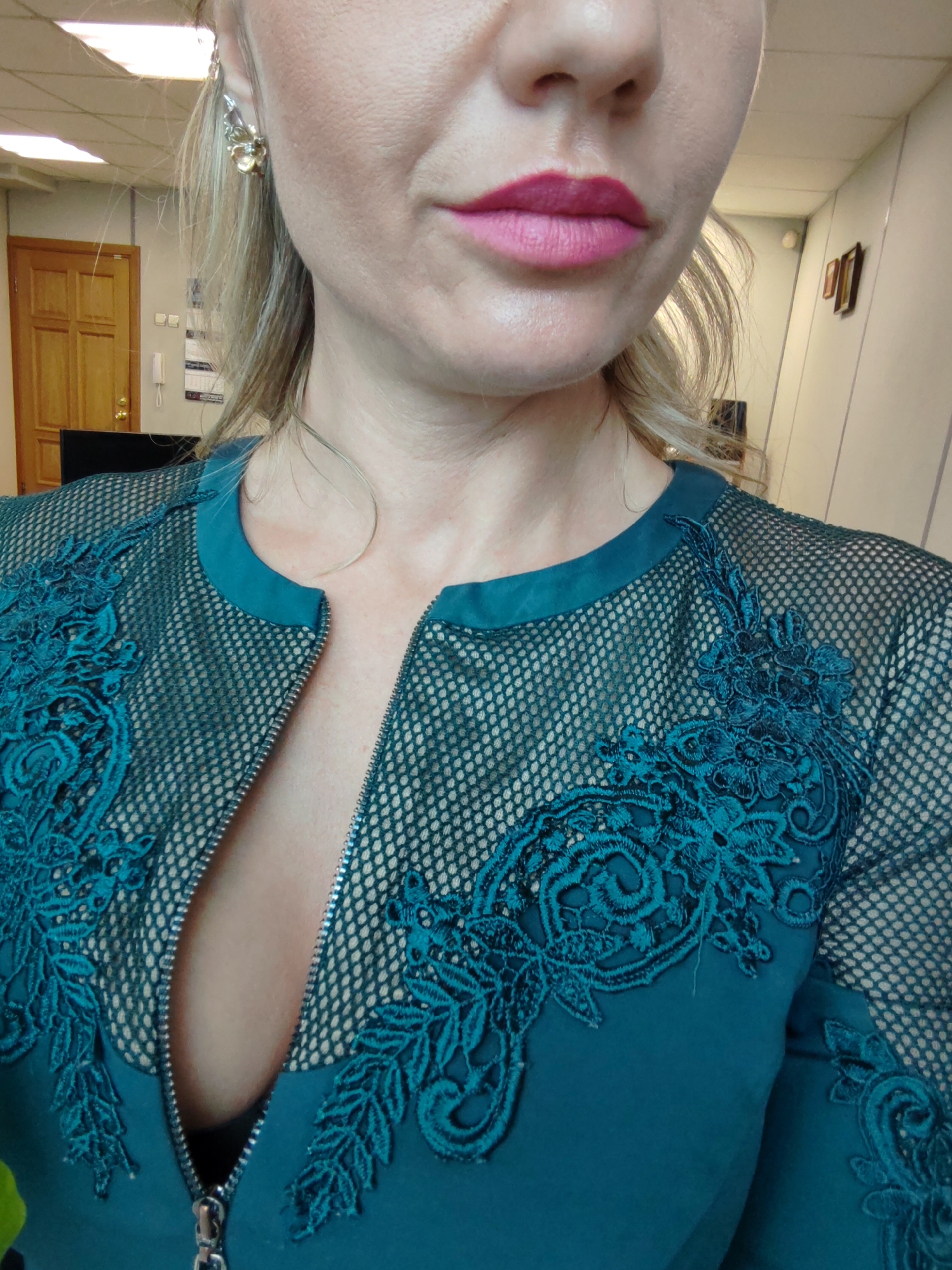 A close-up, waist-up shot shows a fair-skinned woman with blonde hair, wearing a teal jacket with black mesh detailing and teal lace appliques. The jacket has a zipper that is partially open, revealing a hint of her chest. She is wearing bright pink lipstick and a gold and silver floral earring in her left ear. The background is blurred and appears to be an indoor office environment with a wooden door and a wall with two framed pictures.
