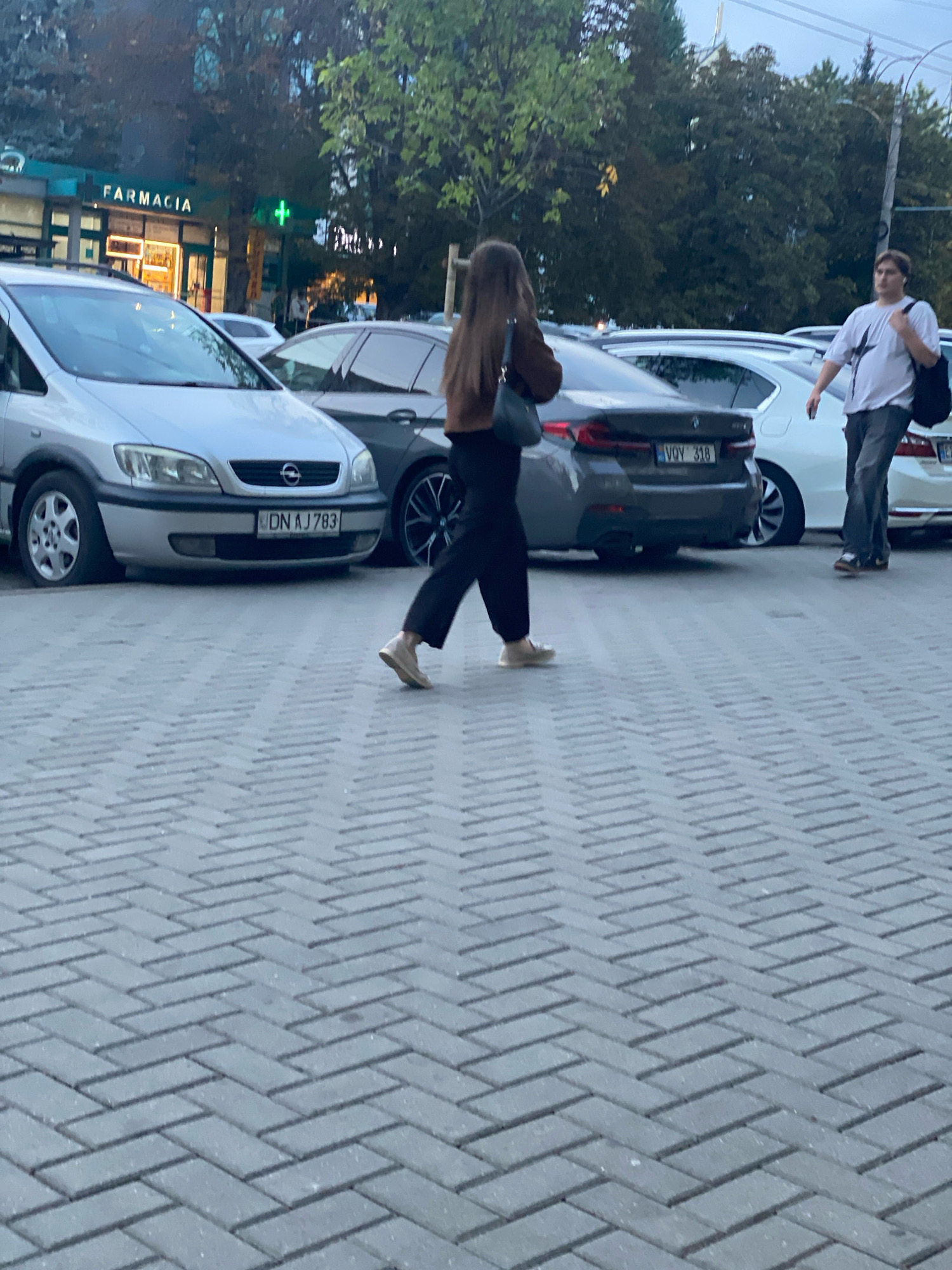 A street scene in Chișinău, Moldova, shows several cars parked on a herringbone brick paved area. In the background, a building with a sign that reads "FARMACIA" and a green cross symbol is visible. Two individuals are walking across the pavement. One person, wearing dark pants and a brown jacket, is walking towards the right of the frame, with their back mostly to the camera. The other person, a male wearing a white t-shirt and dark pants, is walking towards the left, carrying a black backpack over his right shoulder. The scene appears to be outdoors during daylight.