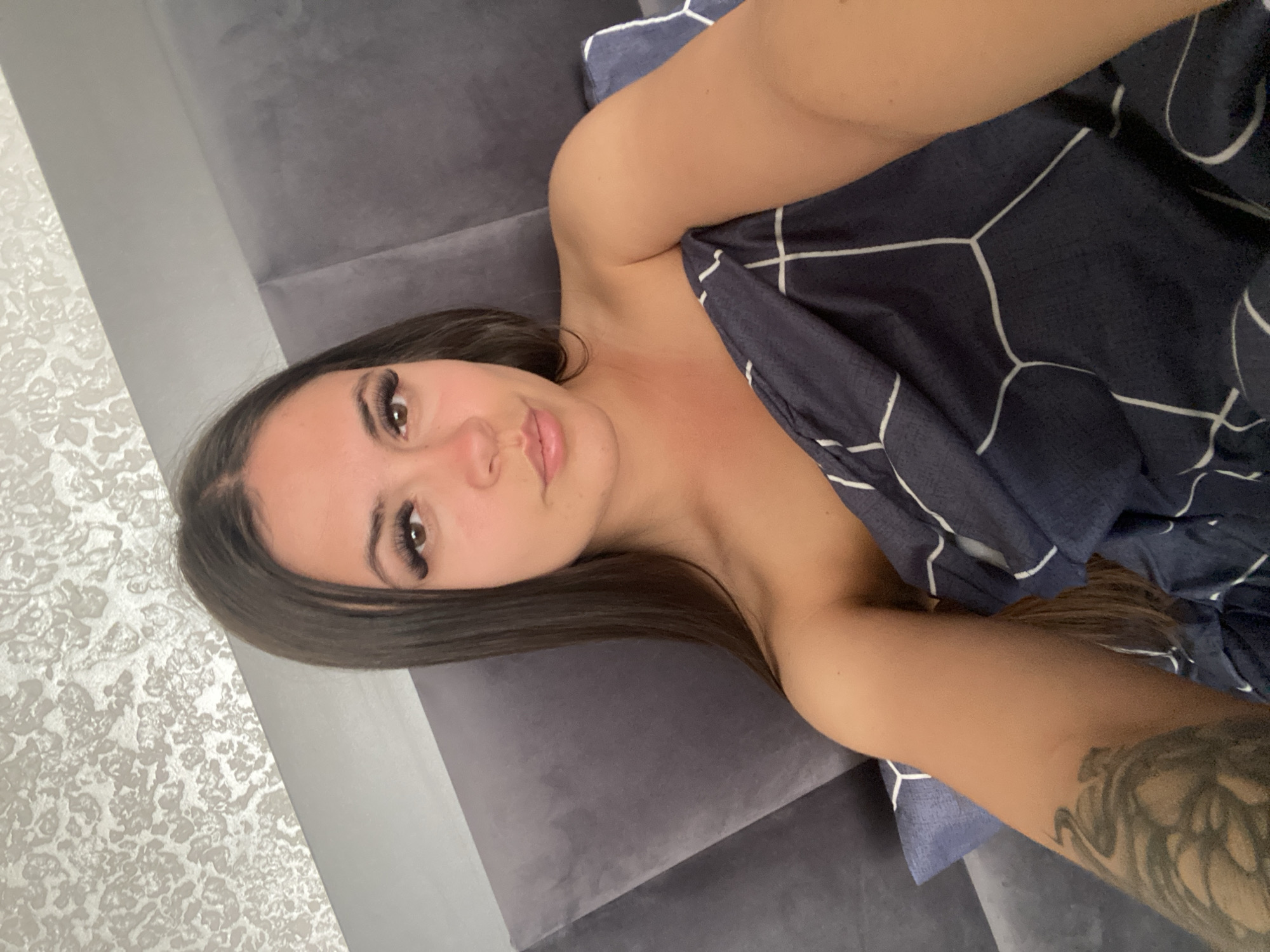 A woman with long dark hair and false eyelashes is lying in bed, covered by a dark blue duvet with a white geometric pattern. She is looking at the camera. Her left arm, which is visible, has a large tattoo on it. The background consists of a grey headboard and a textured wallpaper.
