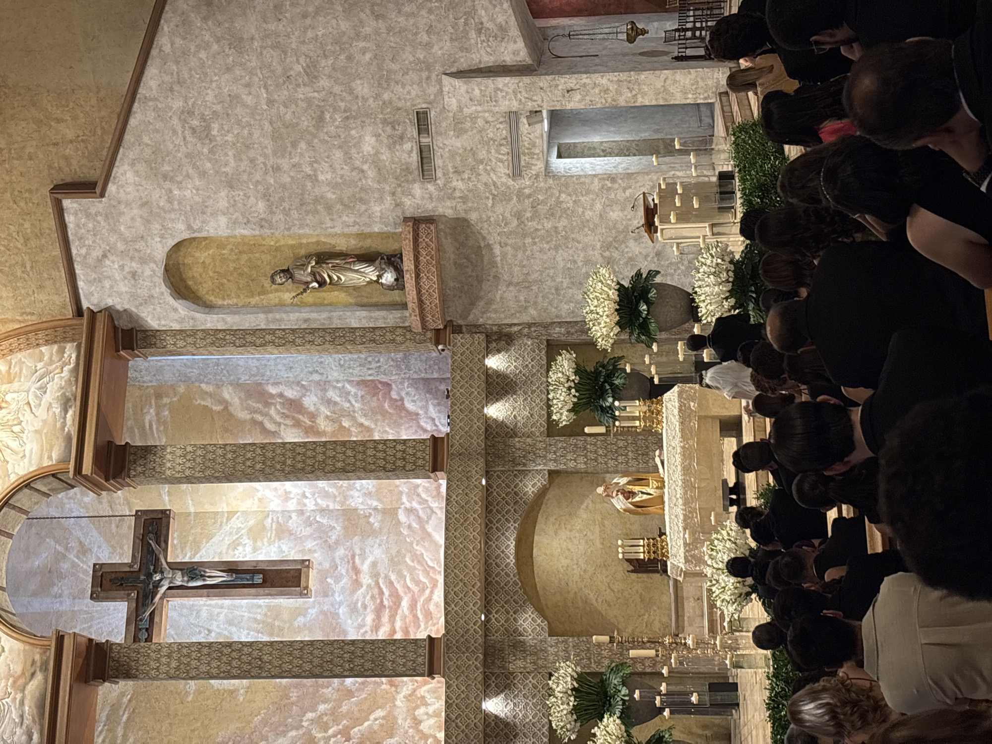 An aerial shot captures an interior scene within a church, likely in San Pedro Garza García, Mexico, during a religious ceremony. In the foreground, a congregation is seated, their backs to the camera, filling the pews. They are dressed in formal attire, with women in dresses and men in suits, suggesting a special occasion such as a wedding or a baptism. The middle ground features an altar area, where a priest in gold vestments is officiating, holding a chalice. The altar is adorned with floral arrangements and candles. The background reveals the ornate architecture of the church, including a large crucifix hanging from the ceiling, columns, and decorative murals depicting clouds. The lighting is dim, with candles providing a soft glow, contributing to a solemn atmosphere.