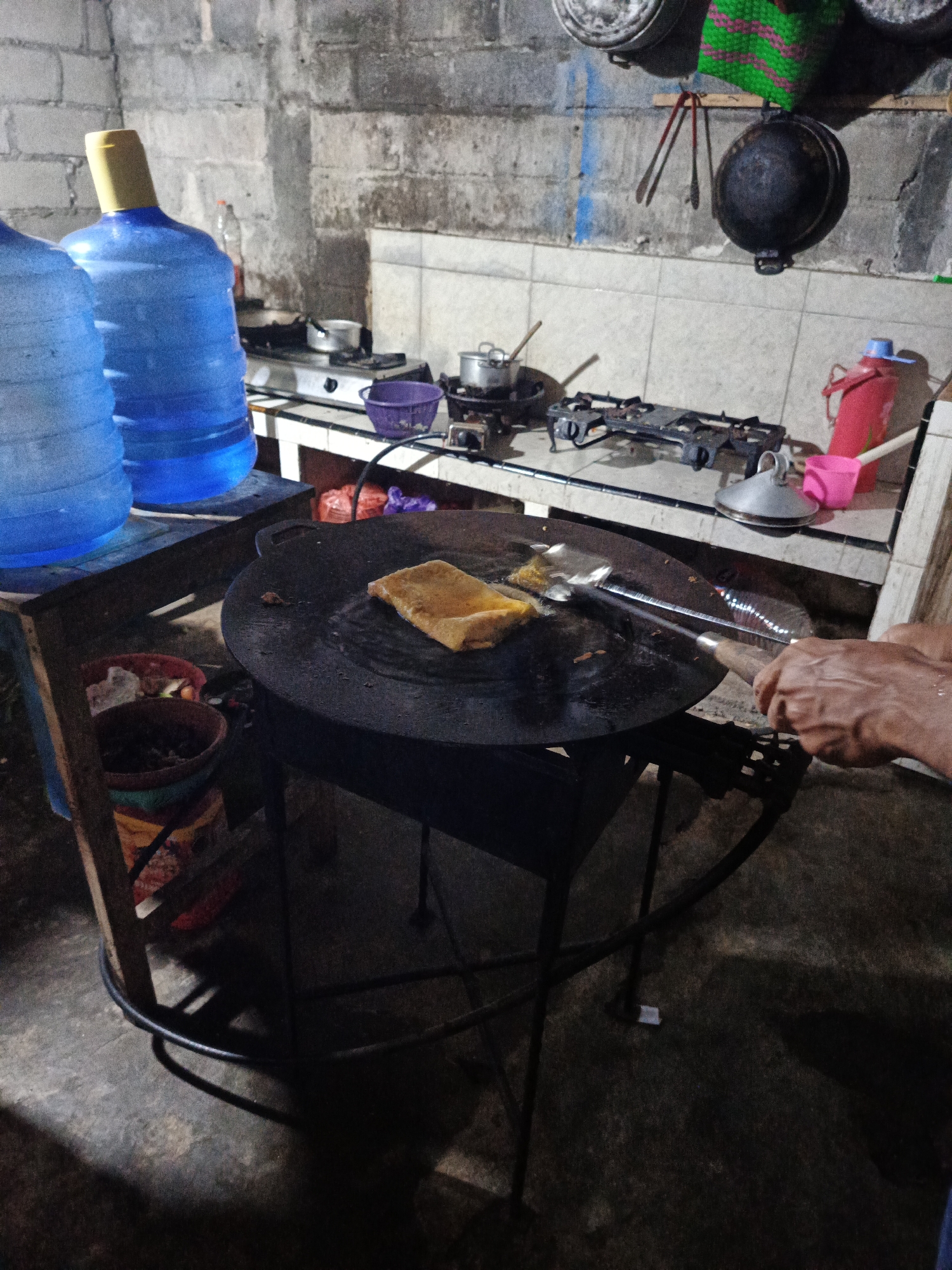 The image depicts a close-up view of a cooking scene likely in Rowokangkung, Indonesia, focusing on a person preparing food. In the foreground, a large, round, dark cooking surface, possibly a wok or griddle, is heated, with oil visibly shimmering. A rectangular food item, appearing golden brown and possibly a type of savory pancake or omelet, is being cooked on this surface. A metal spatula with a wooden handle is actively being used by hands to maneuver the food.
To the left, two large blue water cooler jugs are partially visible, suggesting a domestic or small business setting. In the background, a tiled wall and countertop are visible, with a gas stove, pots, and cooking utensils. A pink plastic pitcher and a purple basket are also present on the counter. The overall environment suggests an outdoor or semi-enclosed cooking area, likely a street food stall or a home kitchen with an outdoor element, given the rough concrete floor and brick wall. The lighting is dim, indicating it might be evening or a poorly lit interior. The presence of the water jugs and cooking setup points towards a functional food preparation area.