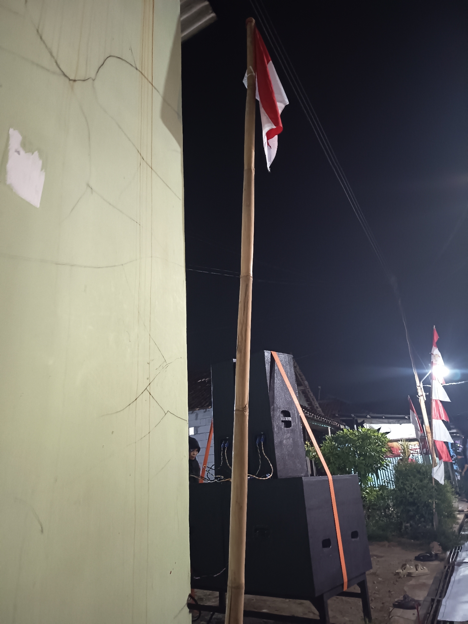 In an outdoor night scene, a light green building wall with visible cracks occupies the left side of the frame. To the right of the wall, a bamboo pole stands upright, adorned with a small Indonesian flag at its peak. Below the flag, a stack of two black speaker boxes is arranged, secured with an orange strap. The speakers are positioned outdoors, with a dark background suggestive of the night sky, punctuated by power lines and distant lights. In the background, residential structures and trees are dimly visible.