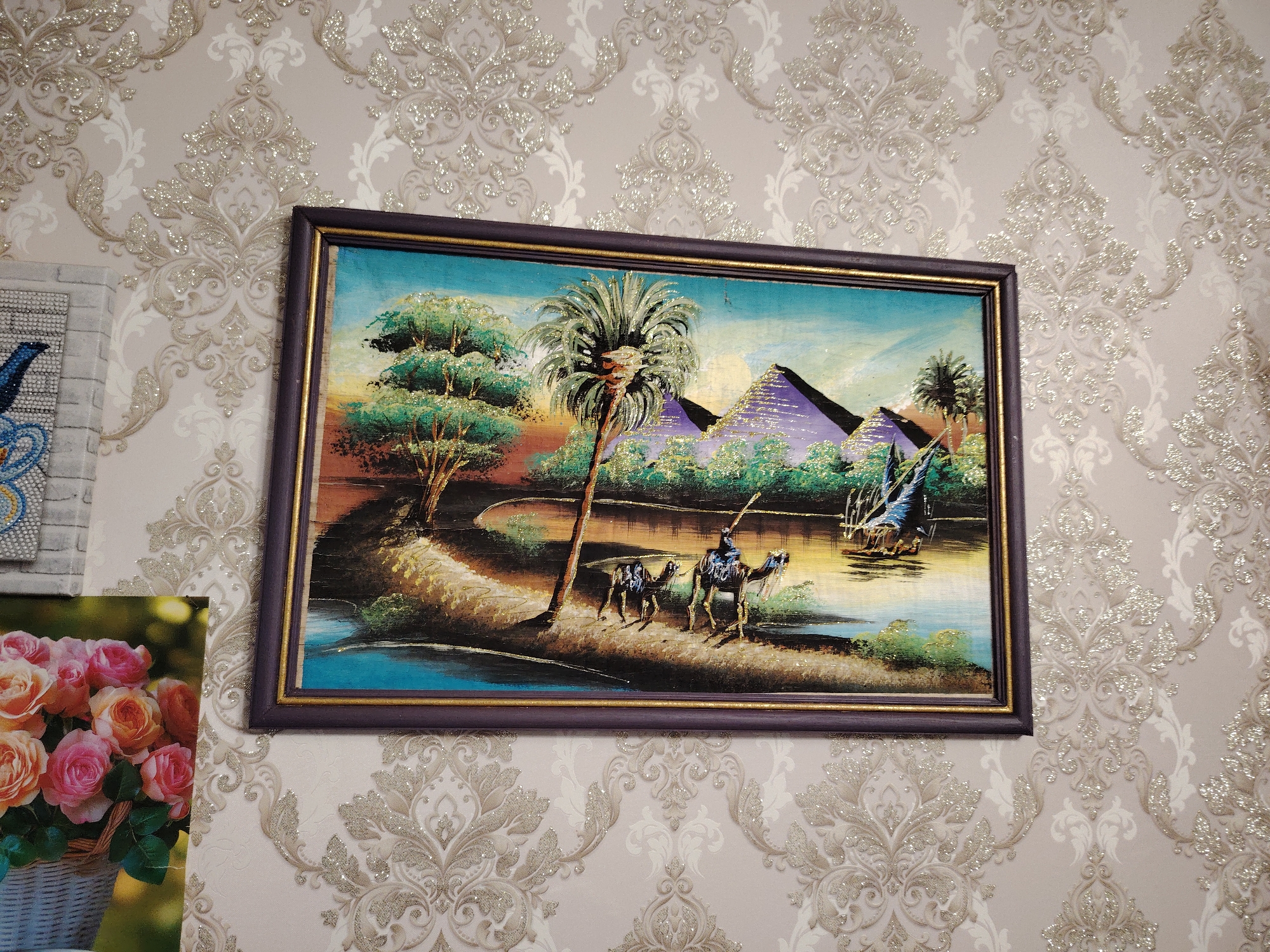 A framed painting hangs on a wallpapered wall. The painting depicts an Egyptian scene with pyramids, palm trees, camels, and a body of water. The frame is dark purple with a thin gold inner border. To the left of the painting, a framed image of roses is partially visible. The wallpaper has a damask pattern with a beige and white color scheme. The scene appears to be indoors. No people are directly visible in the image. There is no indication of the image being in Noviy, Russia.
