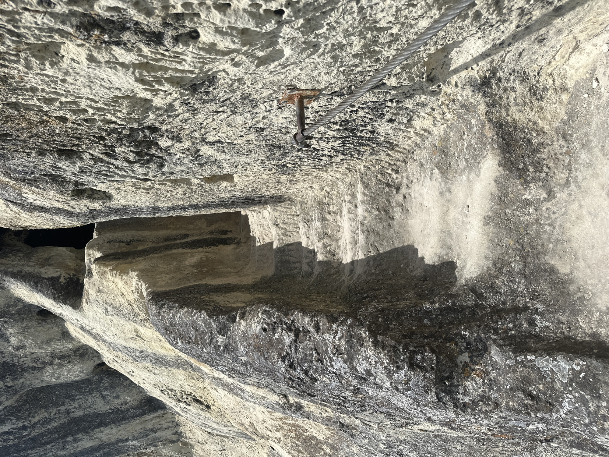 The image depicts a section of a stone structure, possibly a cliff face or the interior of a cave, located in Shumen, Bulgaria. The stone is weathered and appears to be limestone or a similar sedimentary rock. A metal cable runs across the top of the frame, secured to the stone with a rusted metal fixture. The structure features step-like indentations carved into the stone, creating a crude staircase. The lighting is uneven, casting shadows that emphasize the texture and form of the stone.