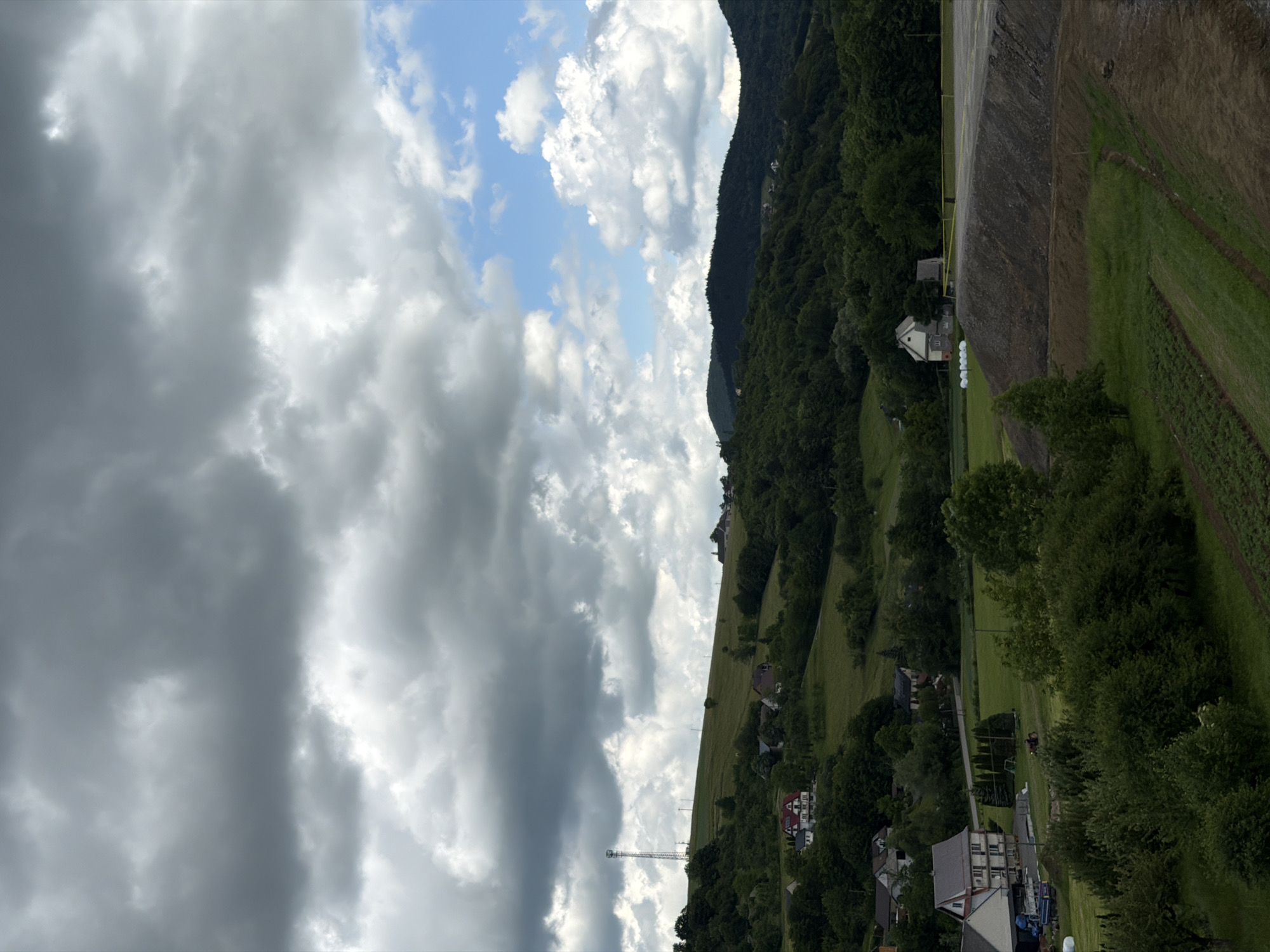 This is a landscape photograph taken on a cloudy day in the city of Janowice, Poland. The image features rolling green hills covered in dense trees and dotted with scattered houses. In the foreground, a construction site is visible, with a large pile of dirt and a paved area. The sky is filled with dramatic, dark clouds interspersed with patches of blue sky and brighter, lighter clouds. A tall telecommunications tower stands on a distant hilltop. There are no people visible in the frame. The scene depicts a rural or semi-rural environment undergoing some form of development or renovation.