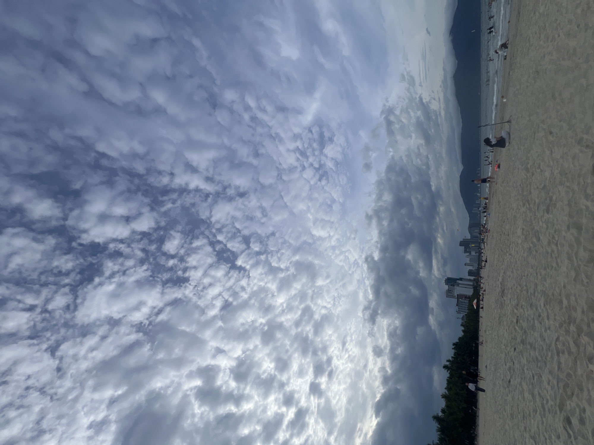 The image depicts a beach scene in the city of Đà Nẵng, Vietnam. The foreground is dominated by a sandy beach with some footprints. In the midground, several people are visible, some walking, others sitting on the sand or near the water. The ocean is calm with some gentle waves. A line of palm trees separates the beach from buildings in the background, which appear to be hotels or apartment complexes, characteristic of a coastal city. Further in the distance, mountains form the horizon. The sky is filled with dramatic clouds, with some areas appearing dark and overcast, while others are lighter, suggesting dynamic weather conditions. The overall atmosphere is that of a late afternoon or early evening on a populated beach.