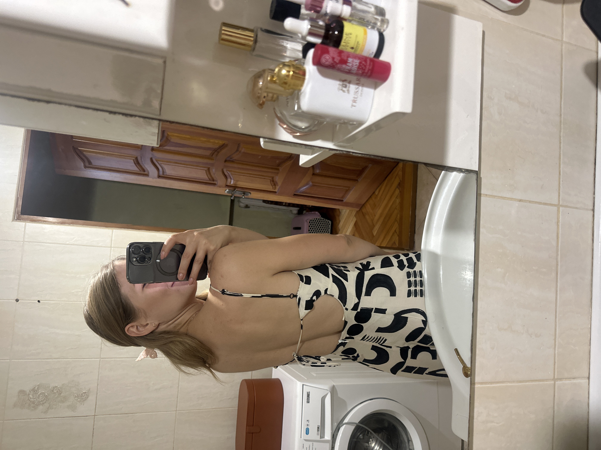 The image captures a woman taking a mirror selfie in a bathroom. She is wearing a black and white patterned dress with spaghetti straps and an open back. Her hair is tied back with a pink accessory. In the background, a washing machine and a brown storage box are visible. A wooden door is also reflected in the mirror, suggesting the bathroom is likely within a residential setting. On a shelf next to the mirror, various cosmetic products, including bottles and tubes, are arranged. The bathroom features tiled walls and a white sink. The overall impression is of a casual, domestic environment.