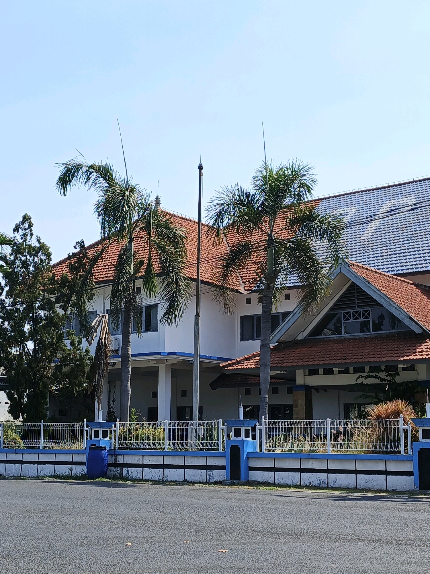 The image depicts a daytime view of a two-story building with a red tile roof, situated behind a white and blue fence. The building has a white facade with visible windows and pillars supporting an upper level. Palm trees are prominently featured in the foreground, with one tree directly in front of the building and another to its left. The fence runs along the front of the property, separating it from a paved road. A blue cylindrical trash bin is visible behind the fence. The sky is clear and blue, suggesting a sunny day. The architecture and vegetation suggest a tropical or subtropical location. The text "Kludan, Indonesia" appears in the metadata associated with this image, indicating the geographical context.