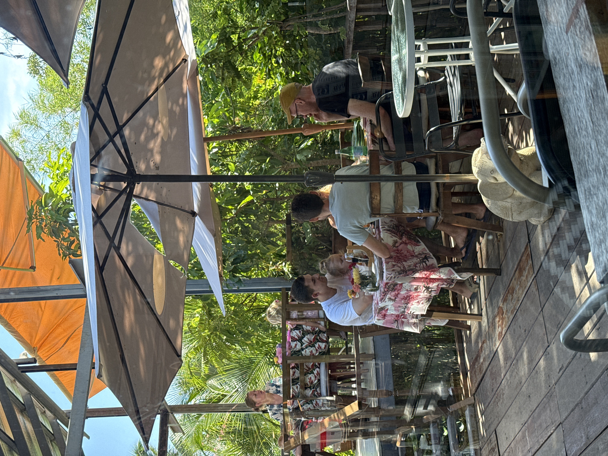 The image depicts an outdoor seating area, likely a restaurant or cafe, located in Ko Samui District, Thailand. The scene is bathed in natural sunlight, filtering through large umbrellas and lush green foliage. Several people are seated at tables, engaged in dining and conversation.
In the foreground, a wooden deck forms the primary surface, with visible grain and some signs of wear. A small elephant statue is positioned on the deck. A metal railing is also prominent in the foreground, suggesting a balcony or a boundary.
Several tables are occupied. One table in the middle ground is covered with a pink and white floral tablecloth. A man in a white t-shirt is seated at this table, holding a young child. Another person, partially visible, is seated to his right. There are flowers and plates on the table.
Further back, another table is set with a floral tablecloth. Two adults are seated here. A woman with blonde hair is looking away from the camera, and a man with a yellow cap is seen from the side, appearing to be speaking.
In the background, the environment is characterized by dense tropical vegetation, including palm trees and other leafy plants. The structures visible include metal beams and orange awnings, suggesting a covered or partially covered outdoor dining space. The overall atmosphere appears relaxed and casual, consistent with a tropical setting.