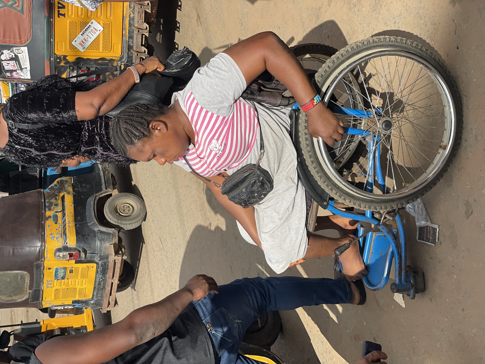 A woman in a wheelchair navigates a busy street in Onitsha, Nigeria. She wears a pink and white striped shirt, gray shorts, and a black purse slung across her shoulder. She is propelling herself forward by gripping the wheels of her blue wheelchair. An unidentified woman, wearing a black, sequined outfit, is assisting her from behind. Adjacent to them is a yellow and black tricycle taxi, common in Nigeria, with the license plate "AKK 170A ANAMBRA." A man wearing blue jeans is positioned in front of the woman in the wheelchair. The scene is set on a paved street under bright sunlight.